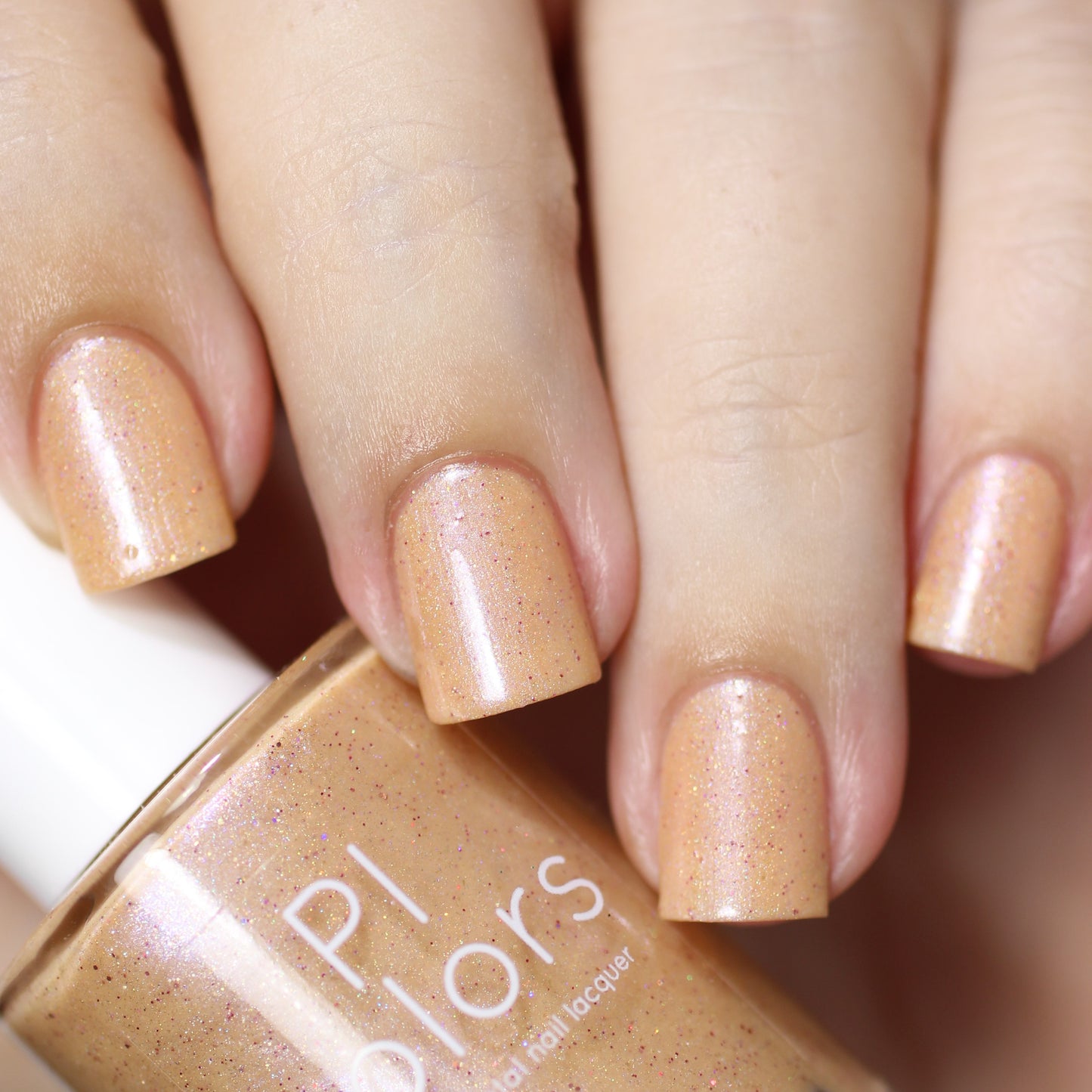 Close-up of a hand displaying nails adorned with Sunrise Tellin.027, a sparkling pale orange nail polish infused with holographic glitter. The polish bottle, featuring a white cap, is held in the hand and showcases the brand name, "PI Colors.