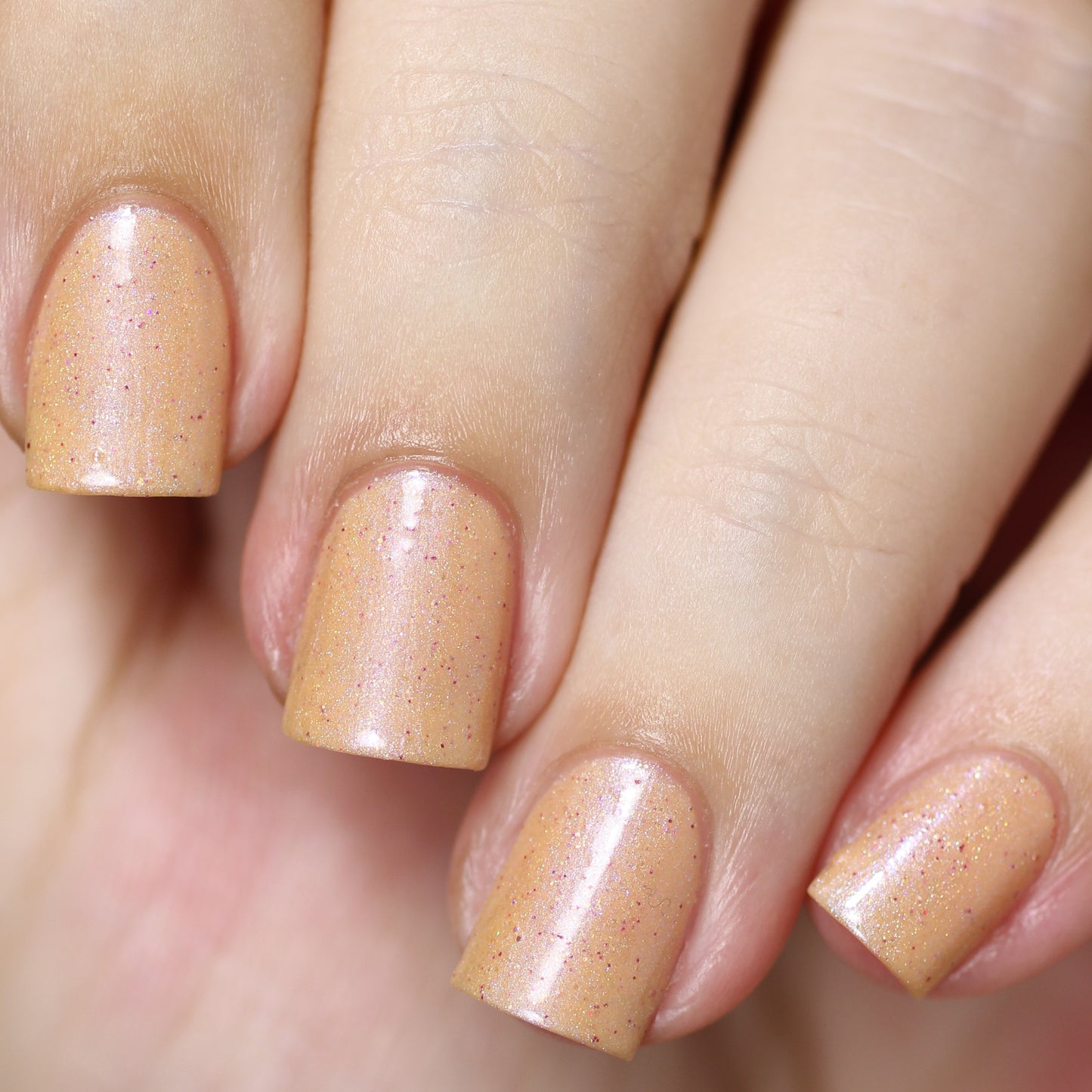 A close-up of a hand showcases a manicure featuring short, square-shaped nails adorned in PI Colors' Sunrise Tellin.027 Pale Orange Nail Polish, a shimmery peachy orange hue with subtle holographic glitter for added sparkle. The light skin tone beautifully complements the delightful nail color.