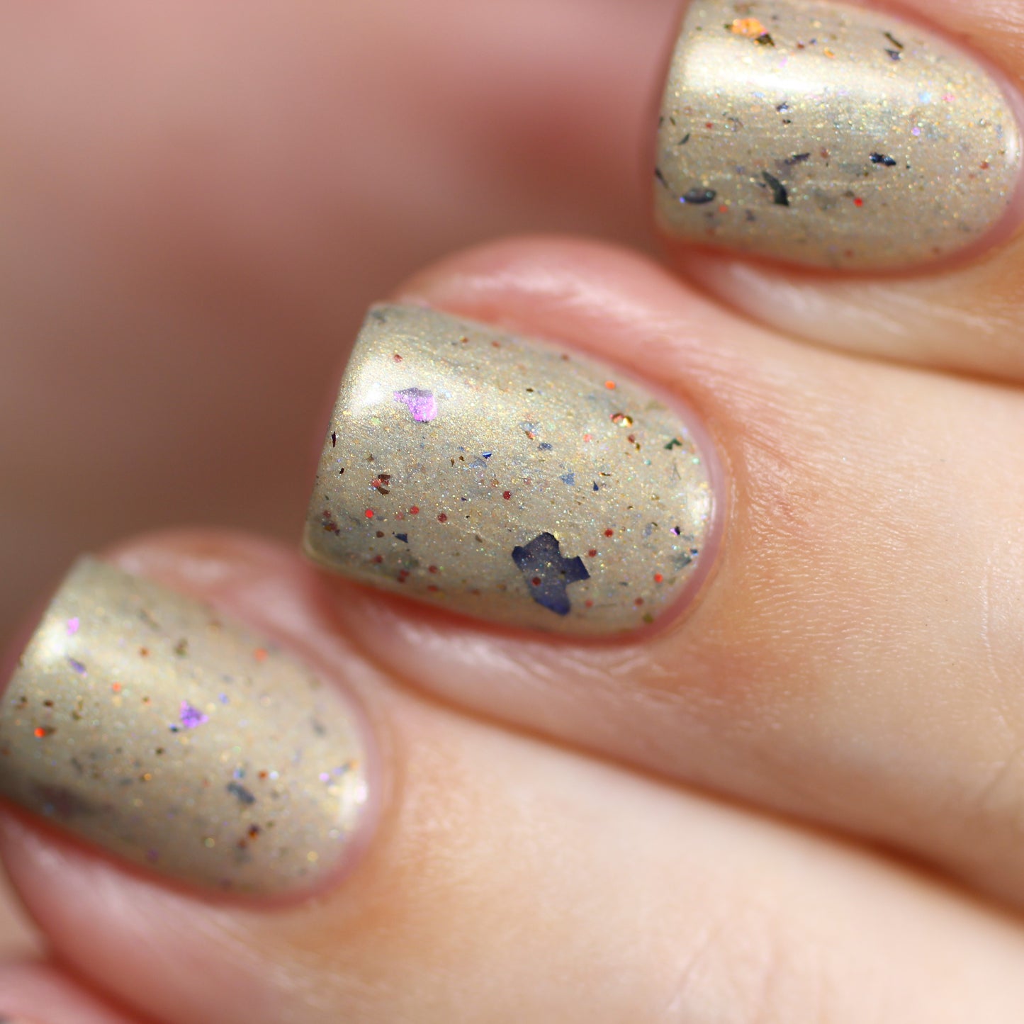 Close-up of fingernails painted with Turtle Dove.102 Taupe Nail Polish by PI Colors, showcasing multicolored speckles and small decorative flakes. The speckles feature shades of orange, blue, and pink, creating a festive and textured appearance with a subtle holographic shimmer for added depth.