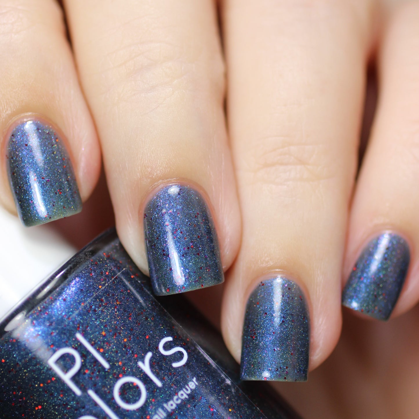 A close-up of manicured fingers with nails painted in a shimmering shade of Winter Berry Blue.001, elevated by holographic powder for an extra touch of sparkle. In one hand, the bottle is held prominently, proudly displaying the "PI Colors" label and emphasizing its exquisite handmade quality.
