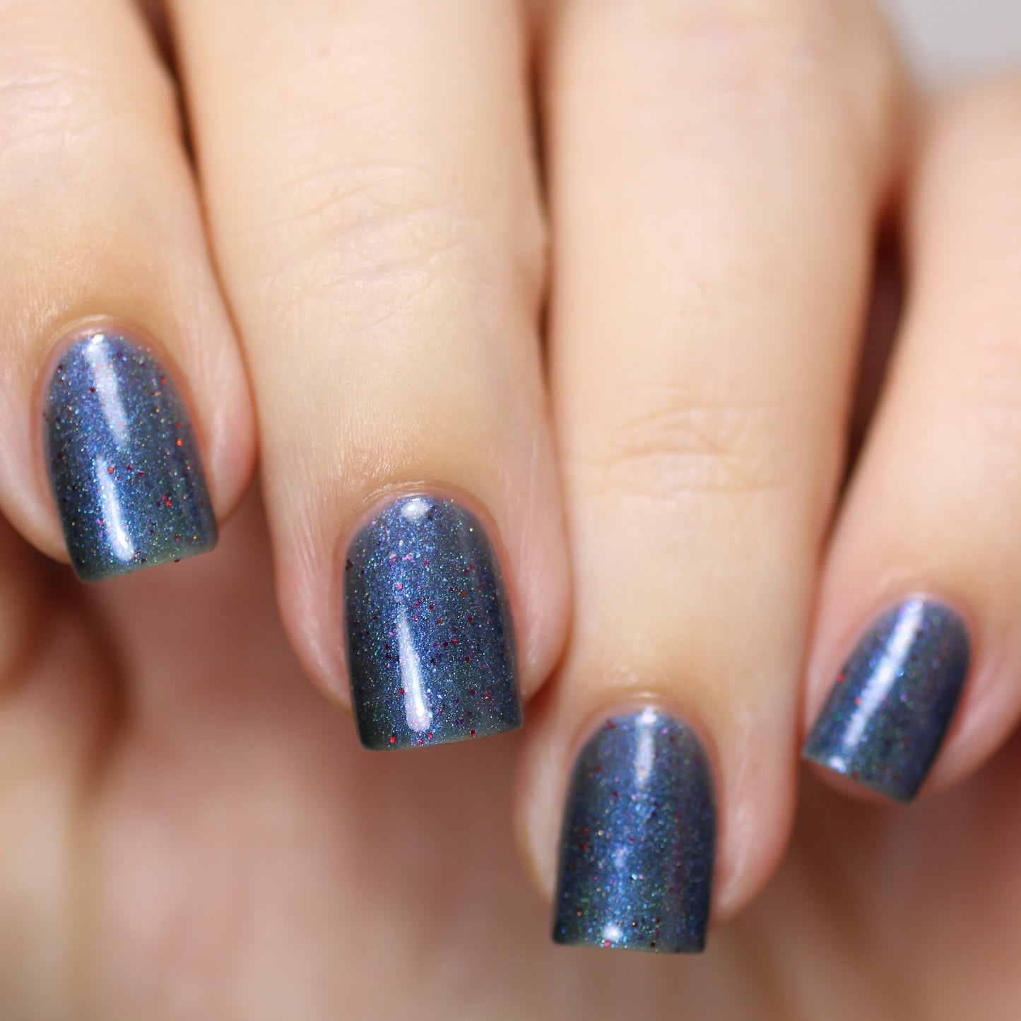 A close-up of a hand displays nails adorned with dark, shimmering polish lightly infused with holographic powder. The short, well-manicured nails radiate elegance with their multicolored sparkles, reflecting the captivating hue of PI Colors' Winter Berry Blue.001 nail polish.