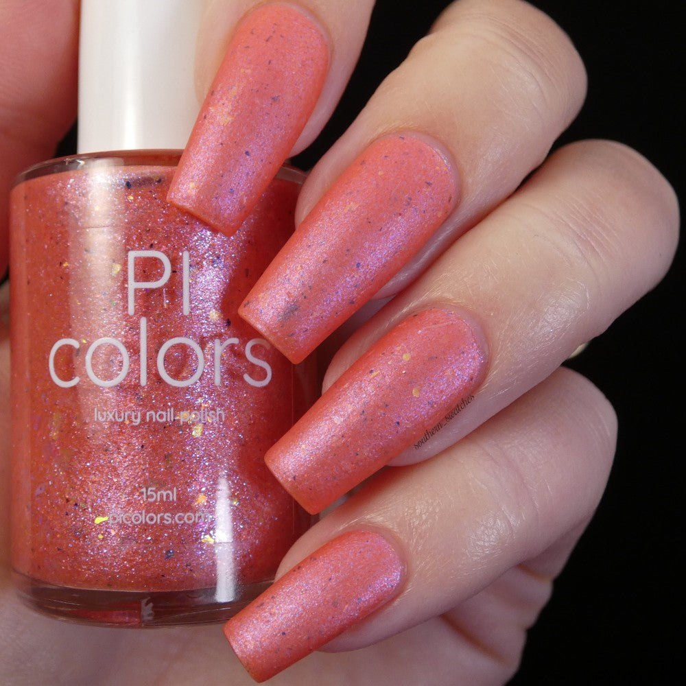 A close-up shot of a hand featuring long nails adorned with a shimmering coral pink polish, holding a custom handmade PI Colors bottle of Lala Ngoxolo Kumkani Coral Nail Polish. The bottle, infused with iridescent rainbow flakes, is nestled between the fingers, showcasing its sparkly texture and vibrant hue.