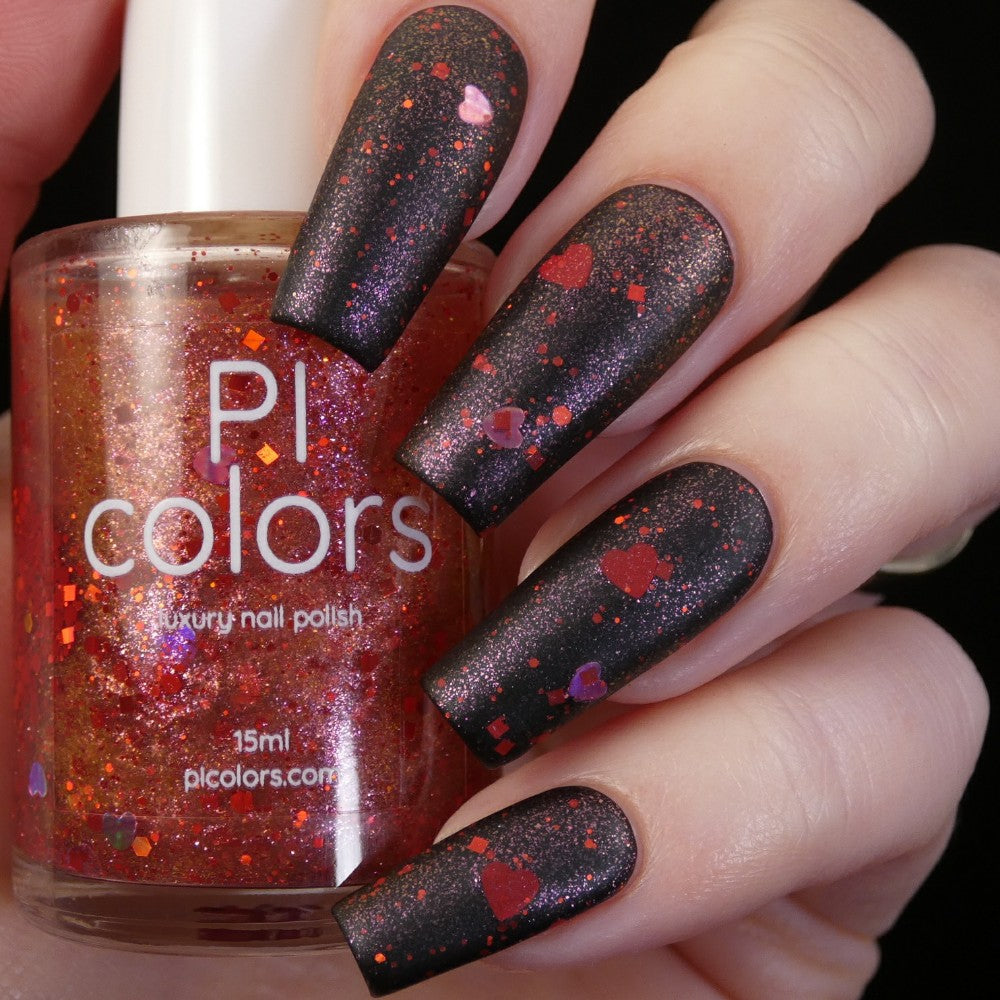 A hand with long nails painted in a dramatic dark polish, decorated with red and orange glitter accents and holographic hearts, holds a bottle of PI Colors Strawberry Heart.208 Red Nail Polish Topper. This 10-free topper enhances the vibrant red-orange hue that perfectly complements the striking nail design.
