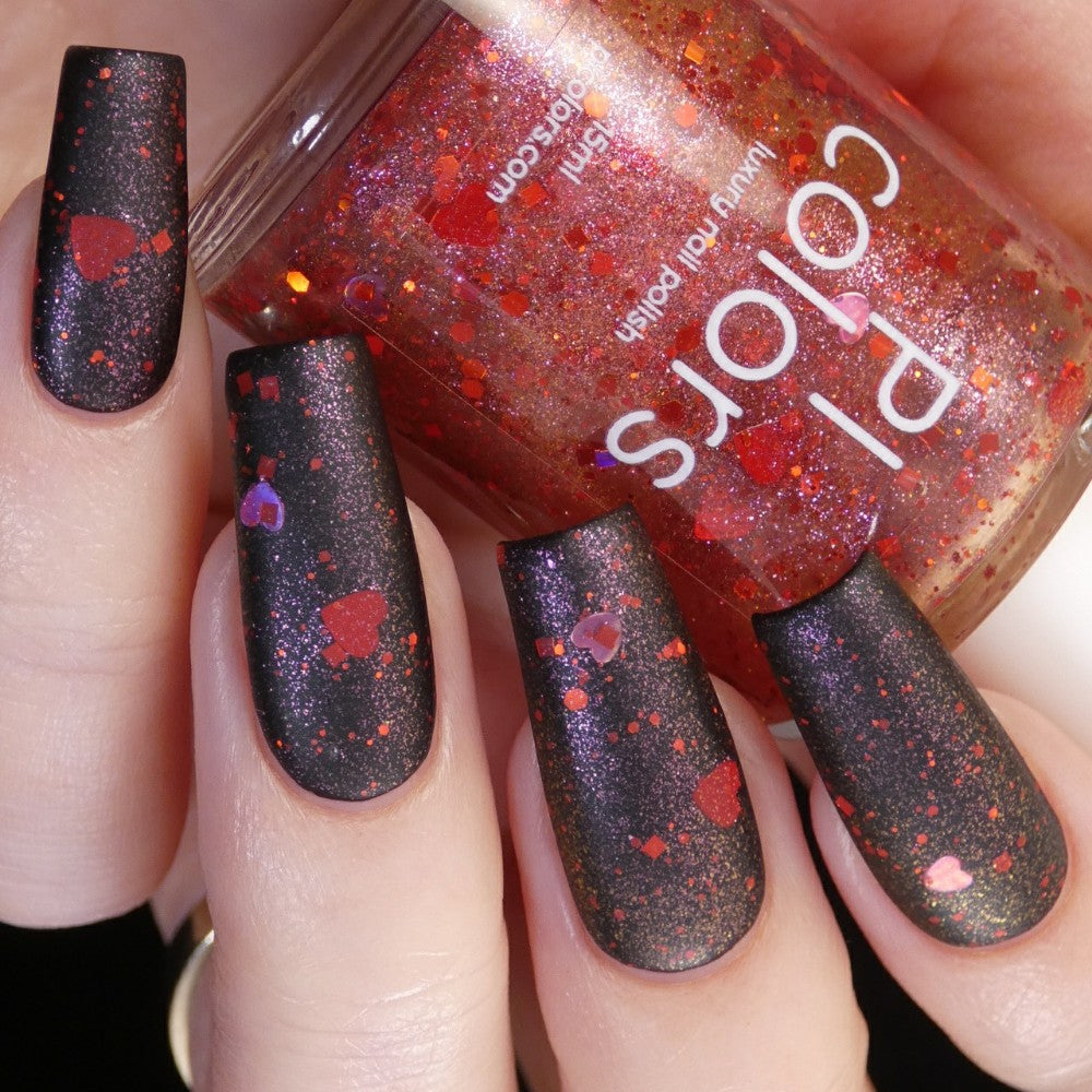 A close-up shot showcases a hand with long, matte black nails embellished with red glitter and holographic heart accents. In the background, a bottle of PI Colors "Strawberry Heart.208 Red Nail Polish Topper" perfectly complements the dazzling design with its matching 10-free red glitter shade.