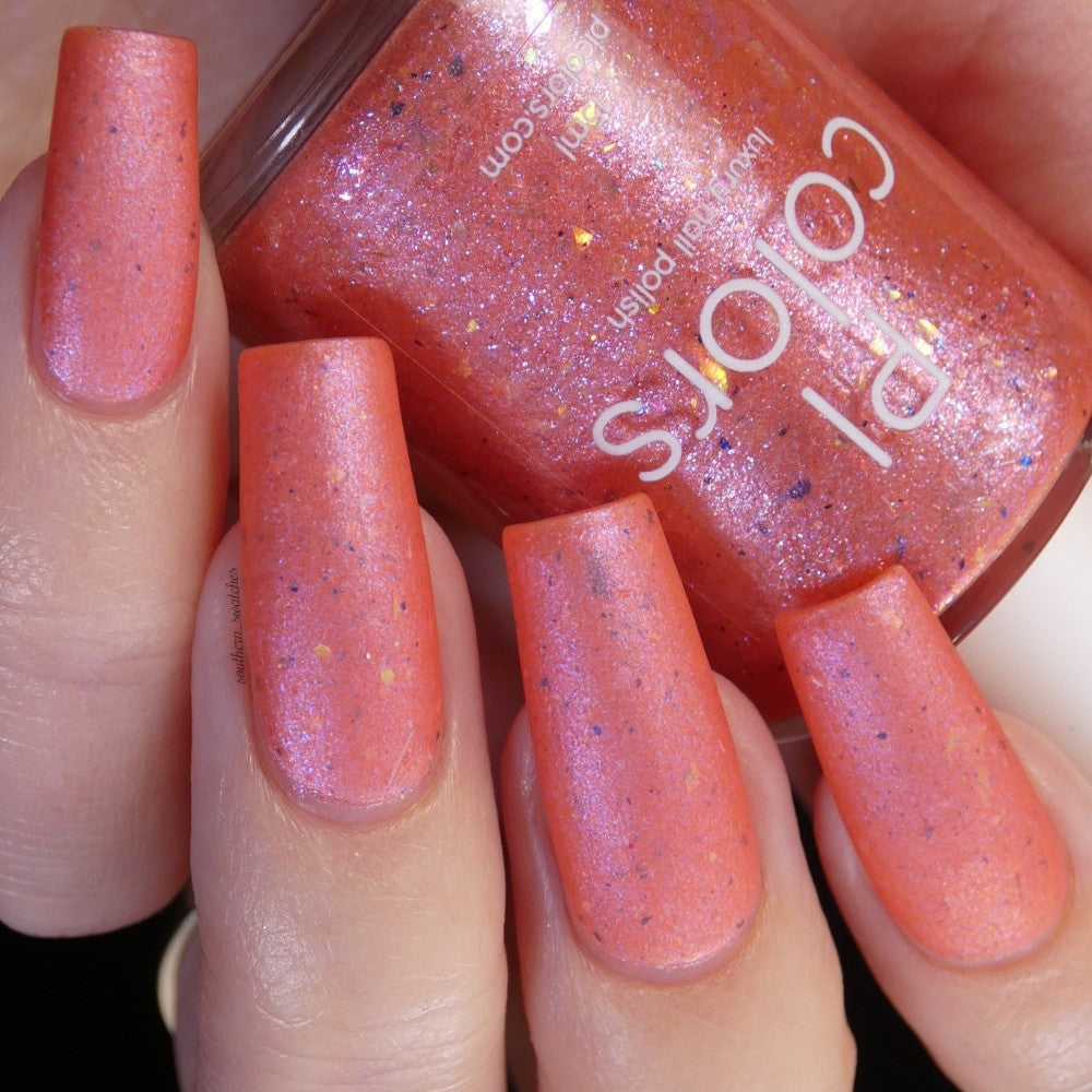 A close-up captures a person's hand featuring long, square-shaped nails adorned with shimmery, peachy coral color using the Lala Ngoxolo Kumkani Coral Nail Polish by PI Colors. A custom handmade bottle of this orange-pink polish, accented with glitter, is elegantly held between fingers. The nails exhibit a glossy, iridescent finish.