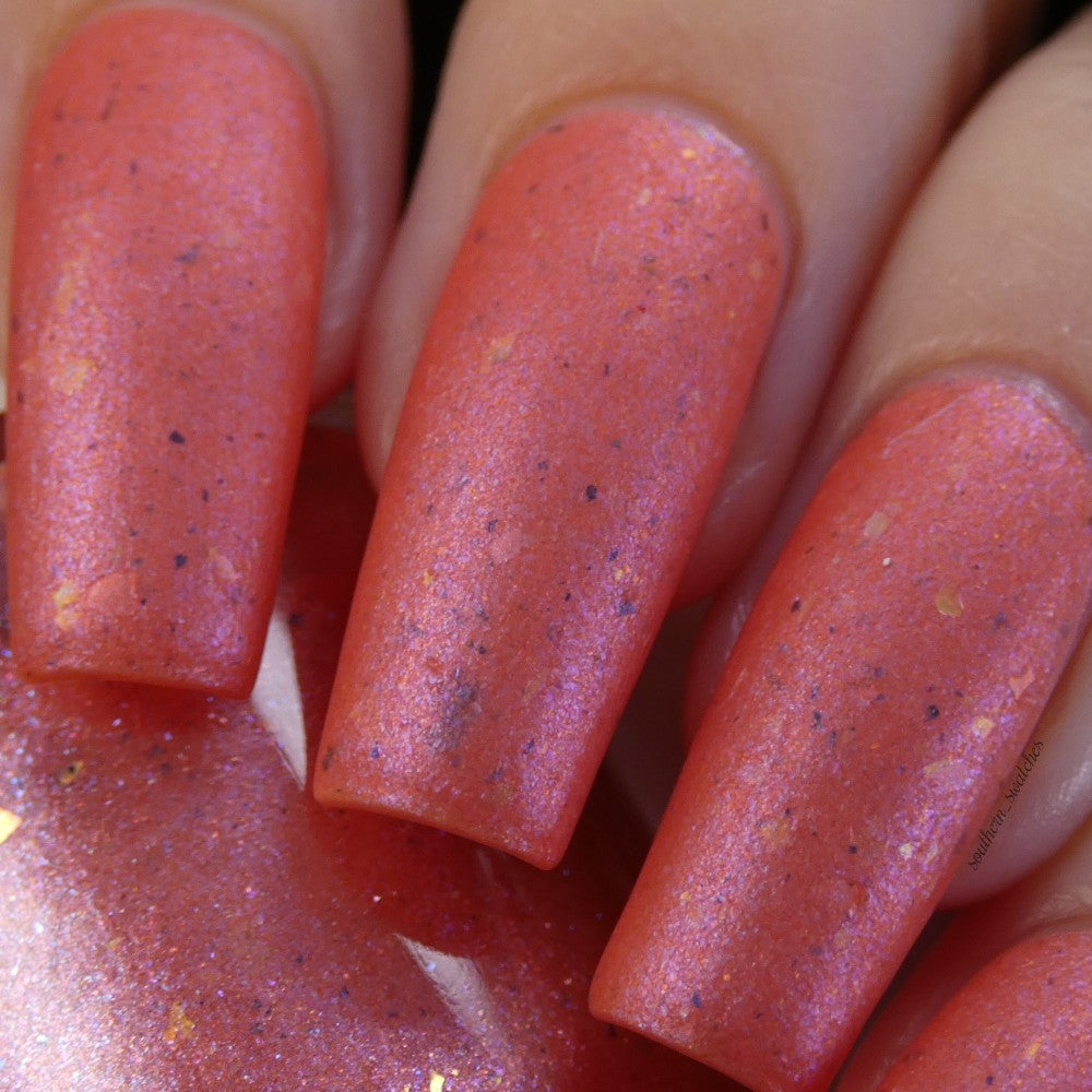 Close-up of nails painted with PI Colors' Lala Ngoxolo Kumkani Coral Nail Polish, showcasing a custom handmade peachy coral shade accented by subtle gold shimmer and black speckles for a textured look. Iridescent rainbow flakes provide depth to the neatly manicured nails, which have a glossy finish.
