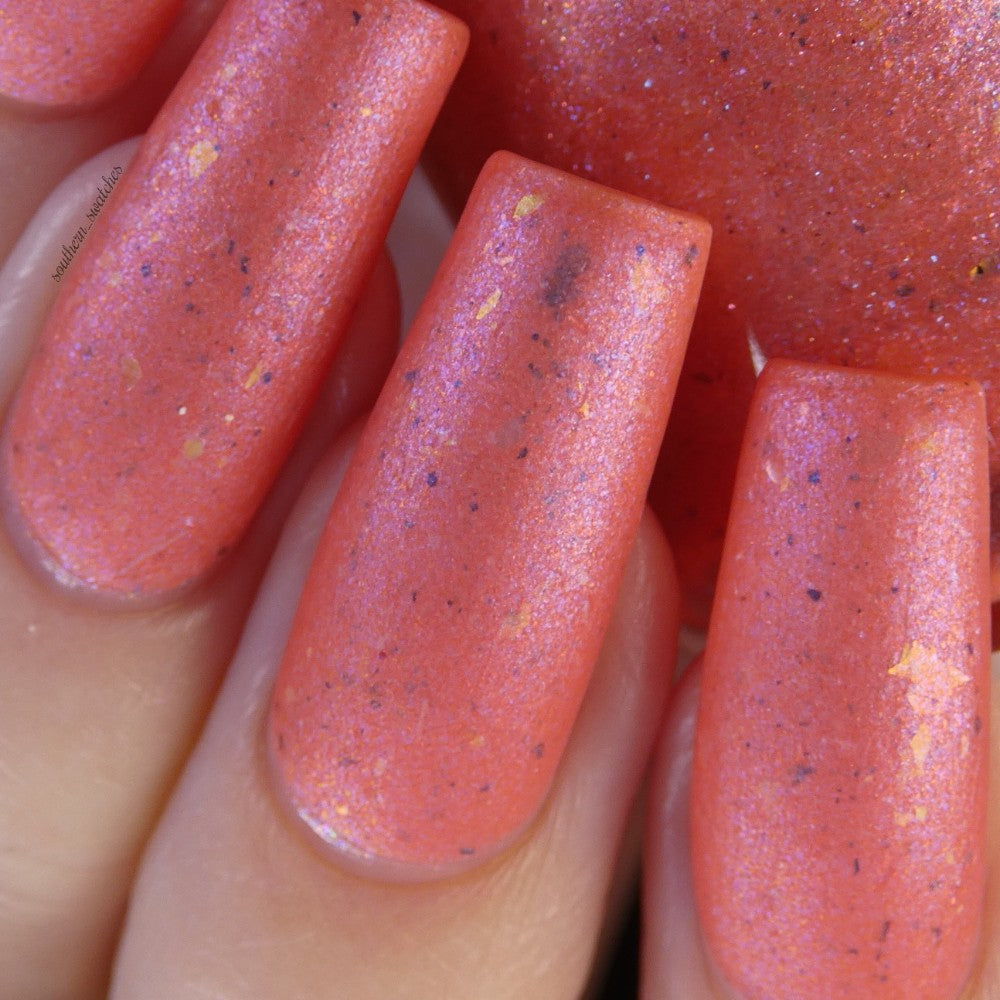 Close-up of a hand showcasing four nails adorned with Lala Ngoxolo Kumkani Coral Nail Polish by PI Colors, featuring a peachy coral hue infused with iridescent rainbow flakes. The polish boasts a shiny, metallic finish with delicate speckles that brilliantly catch the light.