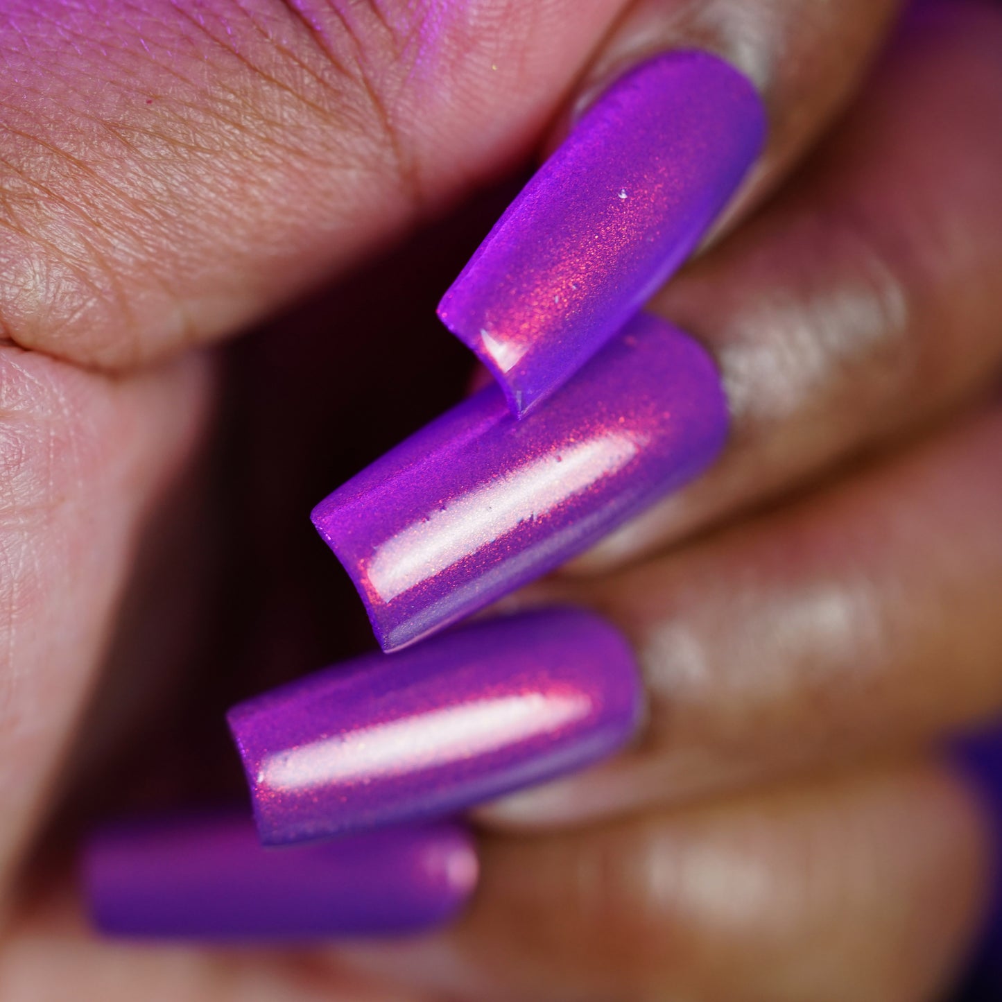 A close-up shows a hand with long, square nails painted in PI Colors’ "Against the Music.100," a handmade luxury nail lacquer in metallic purple with pink undertones. The glossy finish is highlighted as the polished fingers are slightly curled.