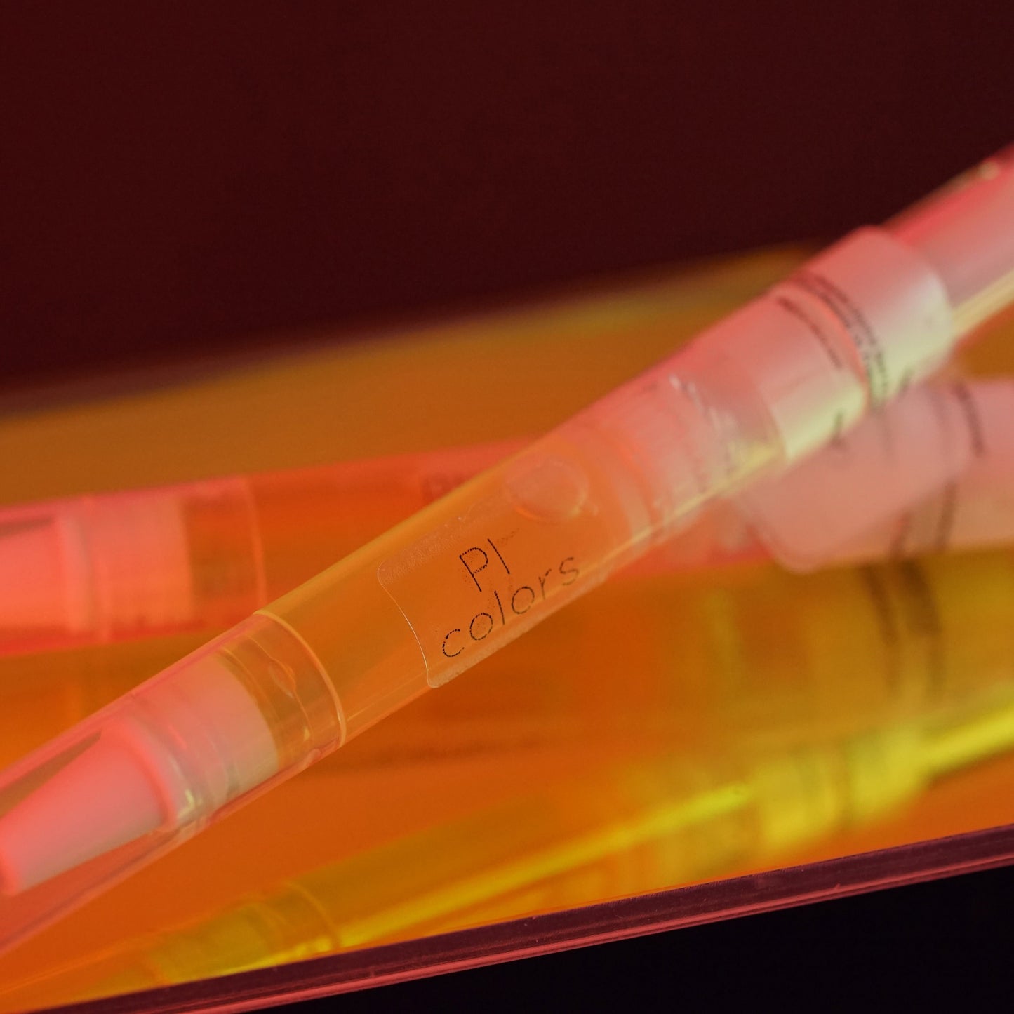 Close-up of a clear pen labeled "PI Colors" resting on a reflective surface amid red and orange hues, emitting the warm and vibrant aroma of natural oil-infused Cuticle Oil Peach Scented.