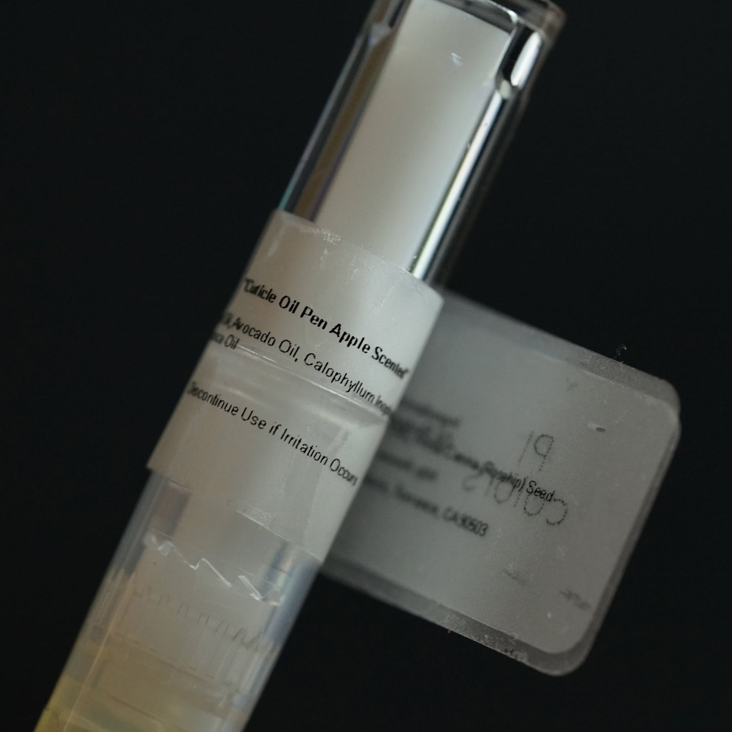 A close-up of PI Colors' Cuticle Oil Apple Scented reveals a partially visible label, showcasing its formulation with jojoba and tamanu oils. The product's clear cap and dispensing mechanism stand out elegantly against a dark background.