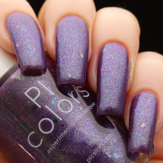 Close-up of hands with long nails painted in Arsu.111 Deep Purple Nail Polish from PI Colors, featuring iridescent flakies. The bottle of nail polish, slightly out of focus, displays a matching hue.