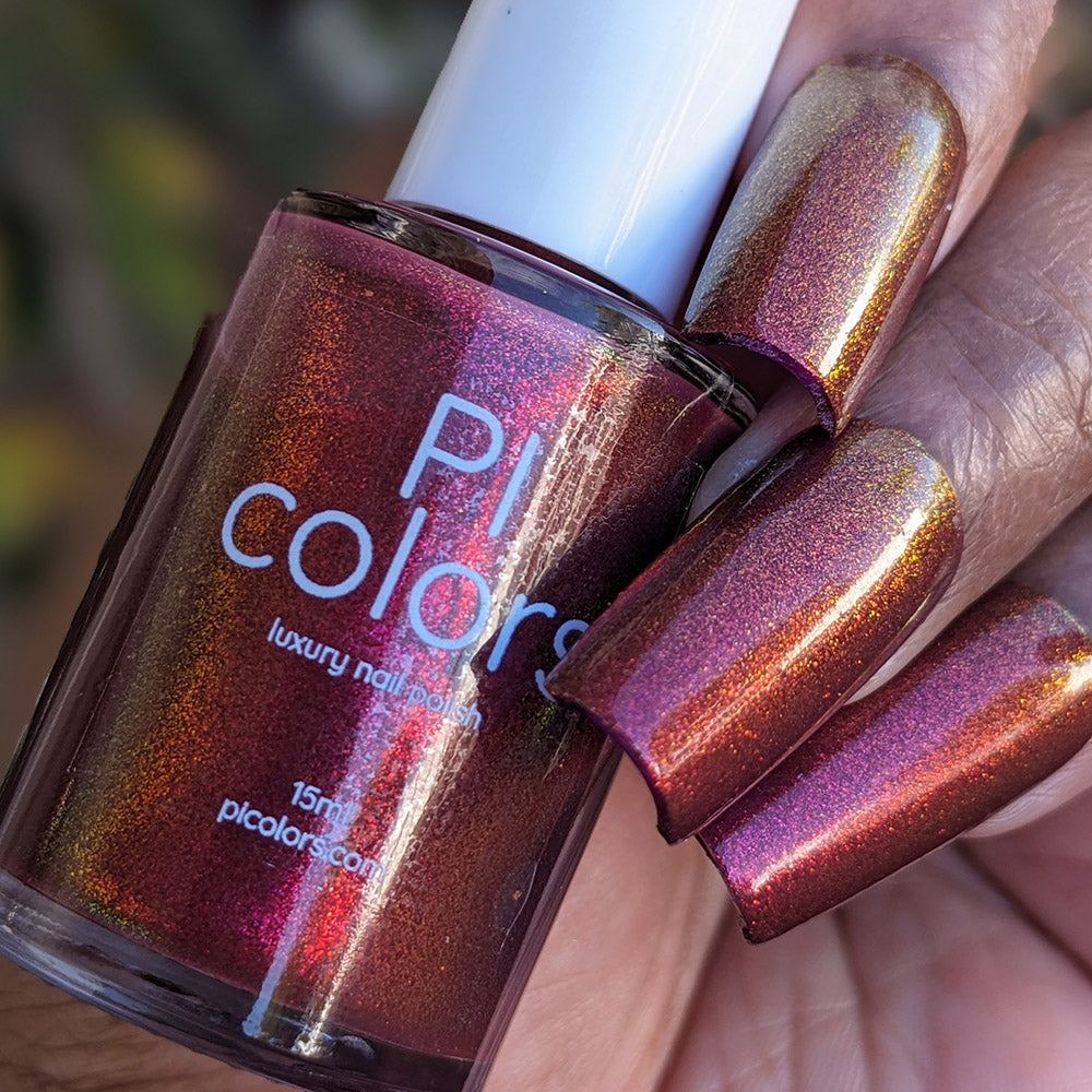 A close-up of a hand with long nails painted in PI Colors "Autumnal Equinox.100," a handmade color-shifting shimmer warm red brown polish, holding a matching bottle labeled "PI Colors luxury nail polish.