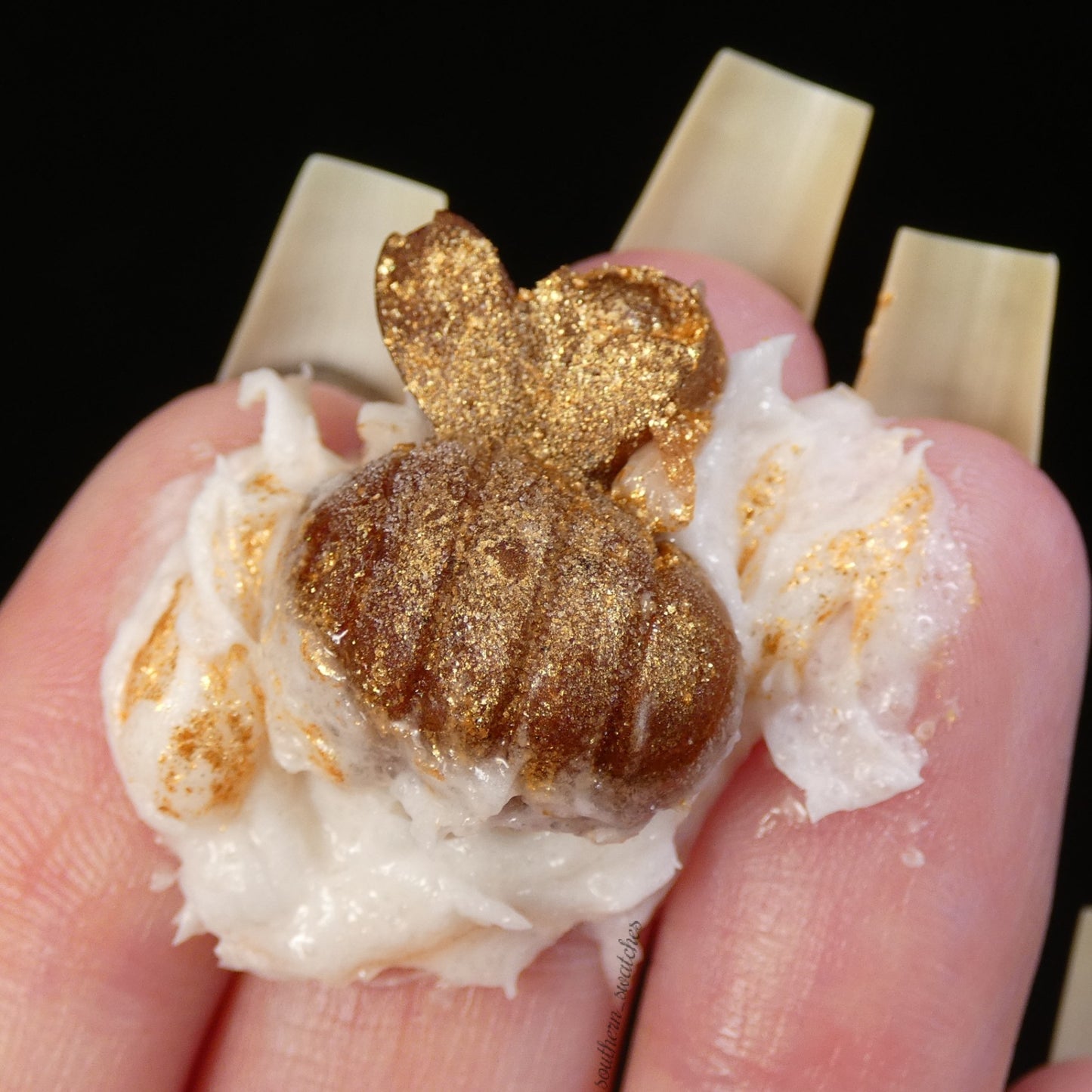 A hand holds a small, glittery golden pumpkin-shaped object atop the Honey Scented Whipped Soap Sugar Scrub by PI Colors. The pumpkin sparkles against the dark background, exuding an air of elegance and charm.