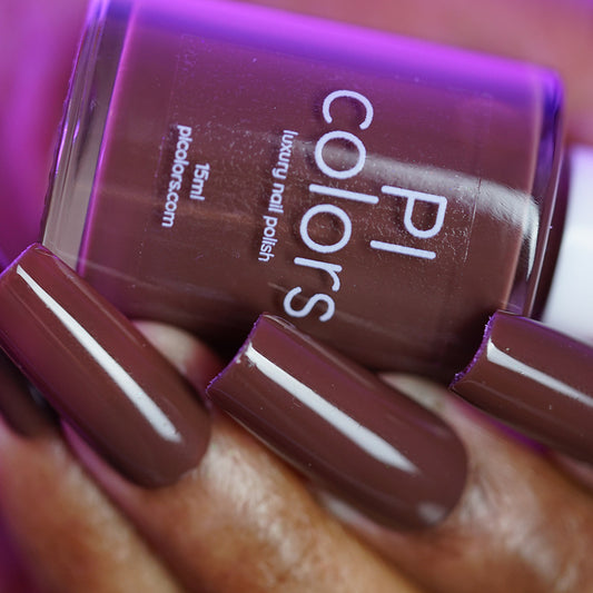A hand with long, glossy nails painted in Chocolate Turtle.411 by PI Colors holds the matching bottle, closely showcased to highlight the smooth brown-mauve creme finish.