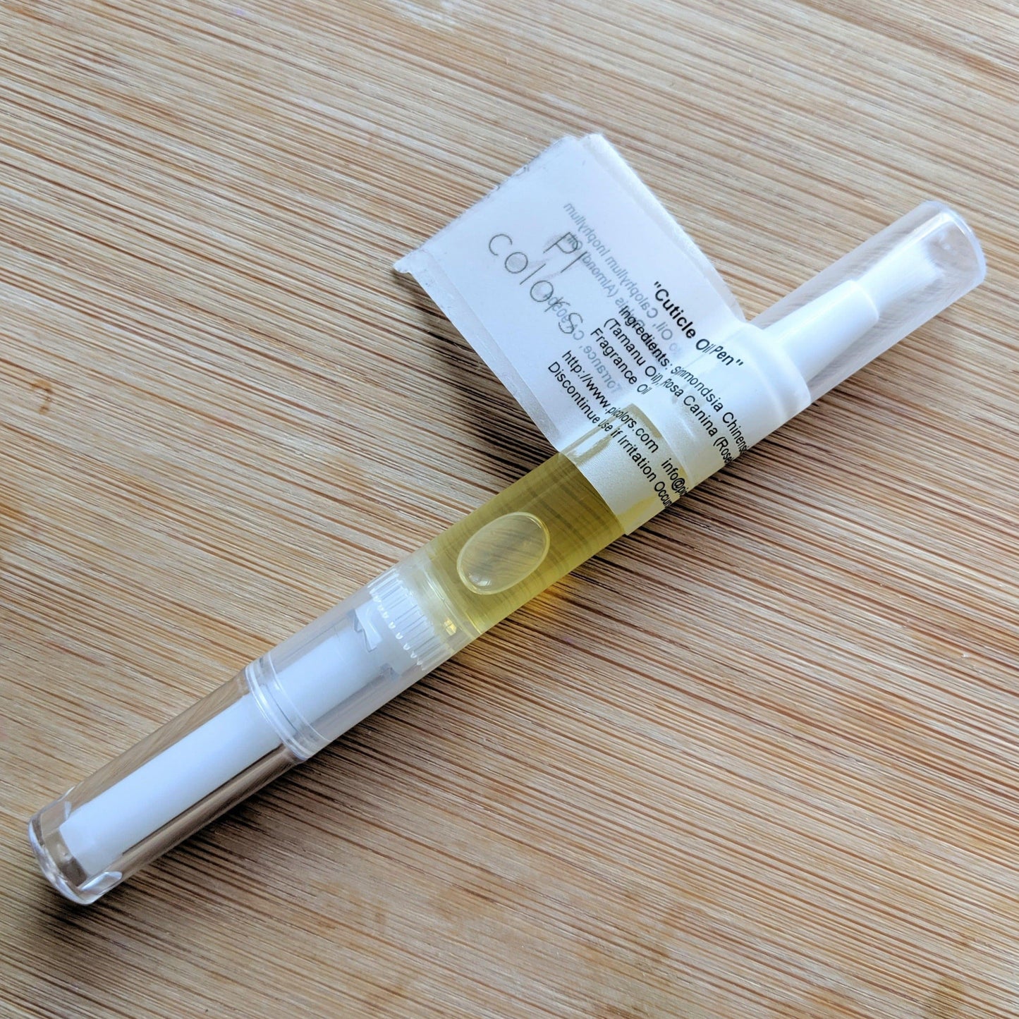 A transparent pen-shaped container with a twist mechanism holds yellow liquid, likely the Cuticle Oil in Blueberry Thyme Scented from PI Colors, on a wooden surface. A label is attached, featuring text and the partially visible brand name.