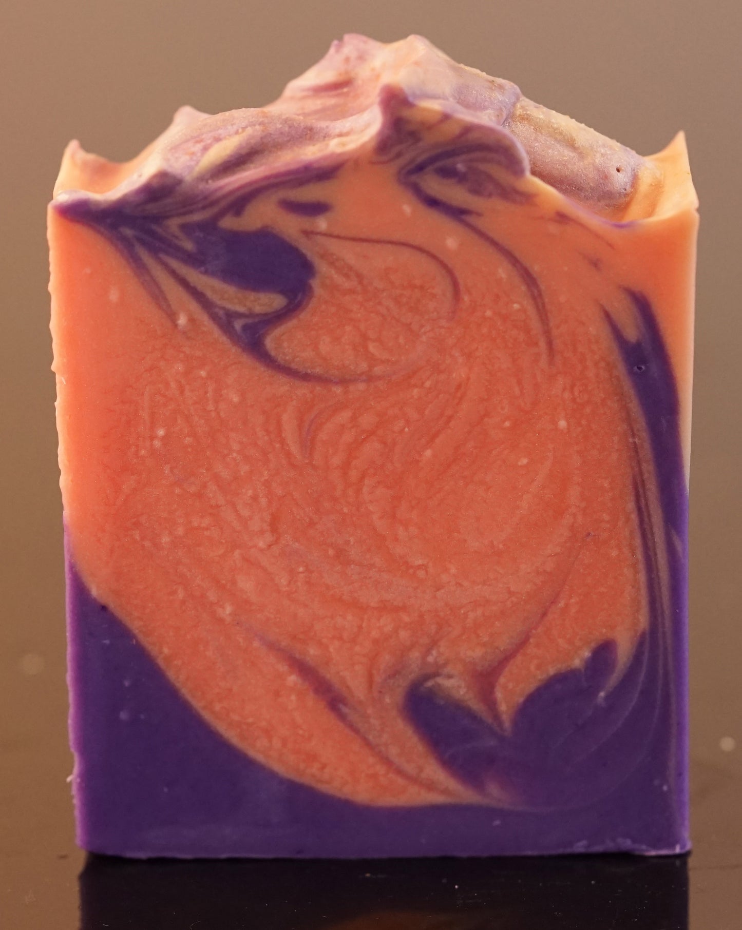 The Autumn Moon Artisan Soap Bar by PI Colors features a rectangular shape with swirled orange and purple patterns. Its top edge is textured and wavy, showcasing intricate marbling that blends colors seamlessly, offering a delightful scented soap crafted from natural ingredients.
