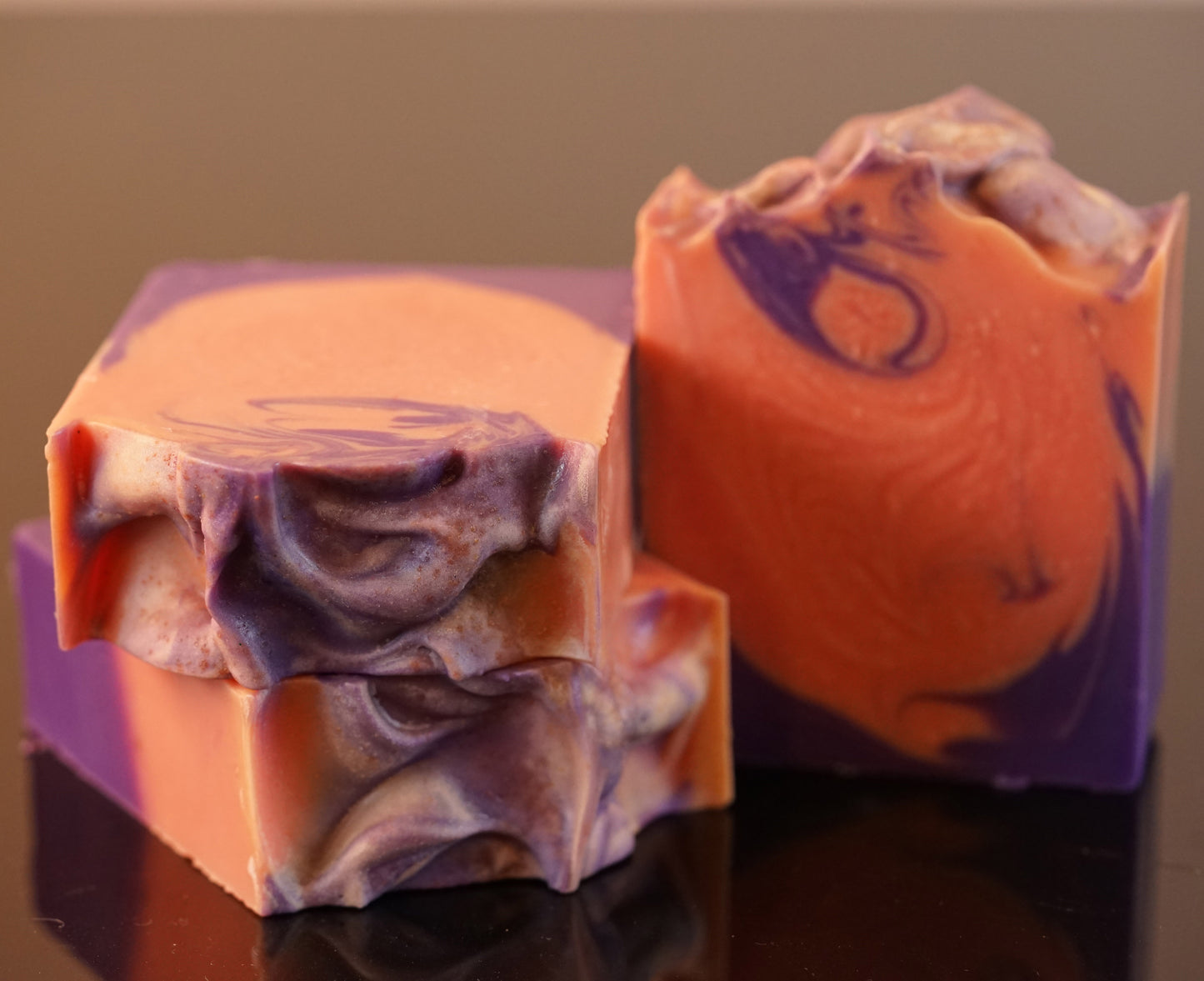 Three Autumn Moon Artisan Soap Bars by PI Colors, made with natural ingredients, display marbled patterns of purple, orange, and white. Their intricate swirls and textured tops catch the light on a glossy black surface, with a hint of spiced orange adding an enticing aroma.