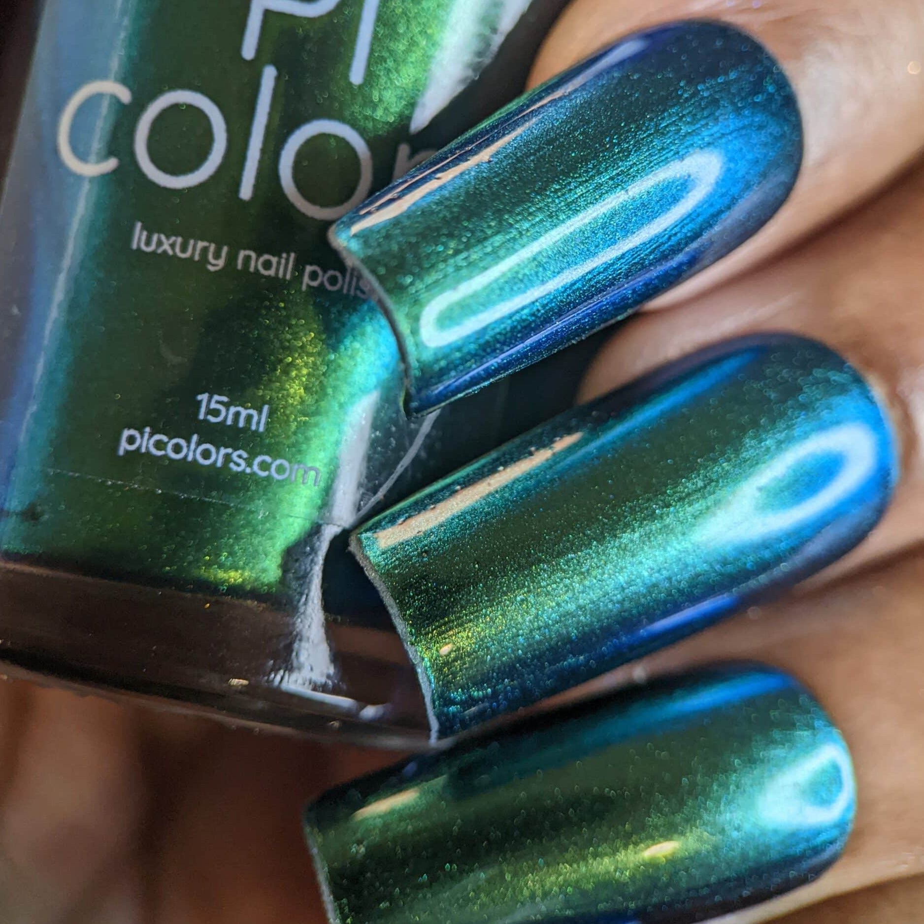 A close-up of a hand showcases nails adorned in a captivating, shimmering multichrome blue-green gradient. In the background, a bottle of PI Colors' Green Glow.209 Green Nail Polish is visible, perfectly matching the nail color.