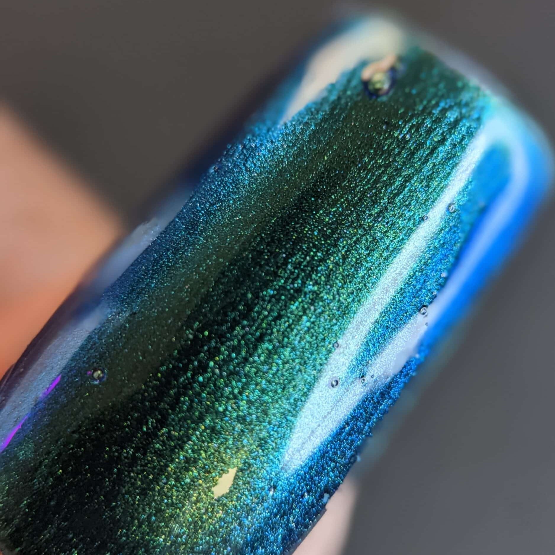 A close-up of a fingernail featuring PI Colors' Green Glow.209 Green Nail Polish, exhibiting a shimmering multichrome finish. The vibrant hues of green, blue, and teal blend smoothly with delicate glitter, reflecting light for a luminous effect.