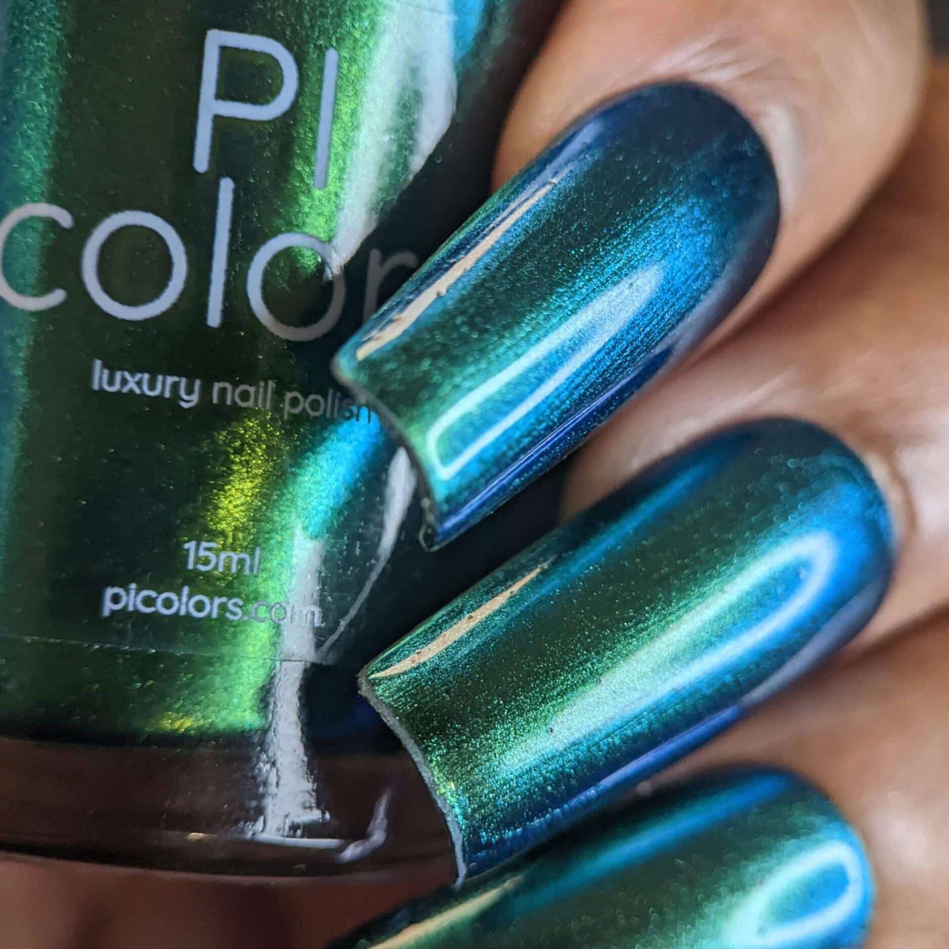 Close-up of a hand with long nails painted in a glossy, multichrome blue-green nail polish. The polish bottle reads "PI Colors" with the label showing 15ml of "Green Glow.209 Green Nail Polish.