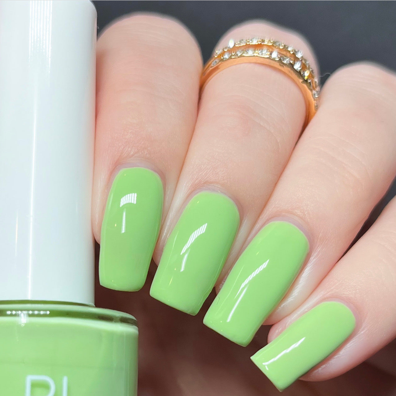 A hand with neatly manicured nails painted in a shiny Pistachio.400 Green Nail Polish by PI Colors holds a bottle of the same hue. The person is adorned with a gold ring accented with small crystals, adding an elegant touch to the vibrant look.