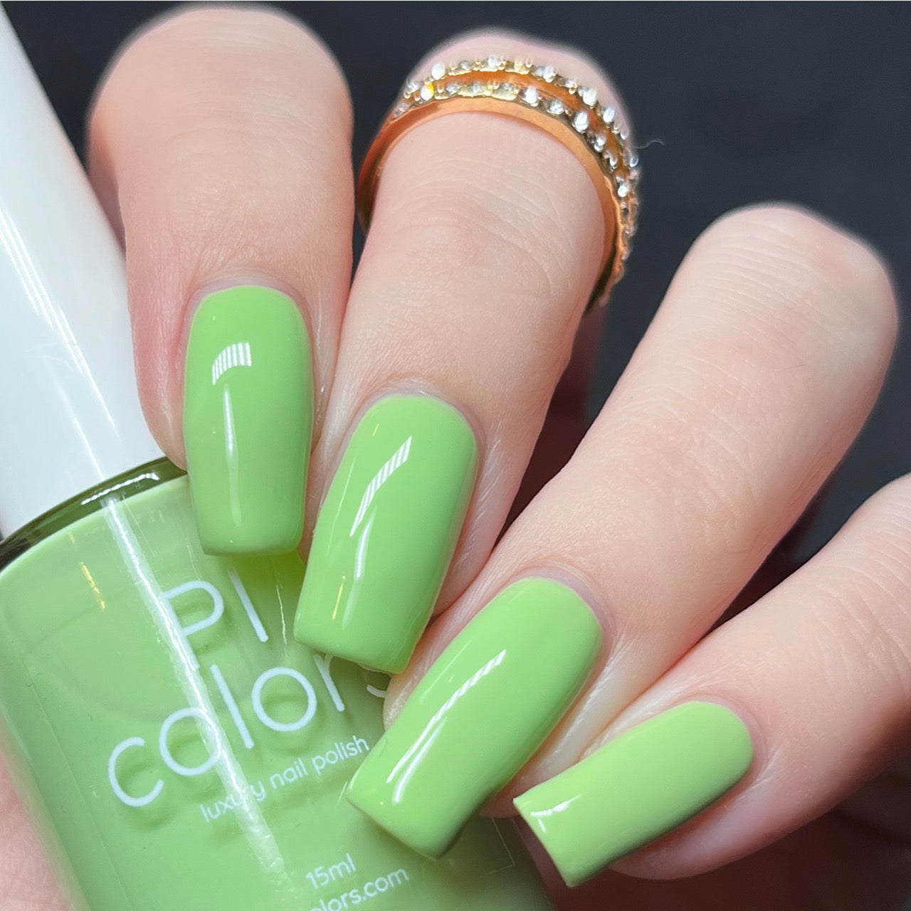 A hand with long nails glistens in a vibrant shade of Pistachio.400 Green Nail Polish. The thumb grasps a bottle bearing the PI Colors label, perfectly matching the polish on the nails. Adorned with a gold ring set with small stones, the person's nails exhibit a glossy creme finish.