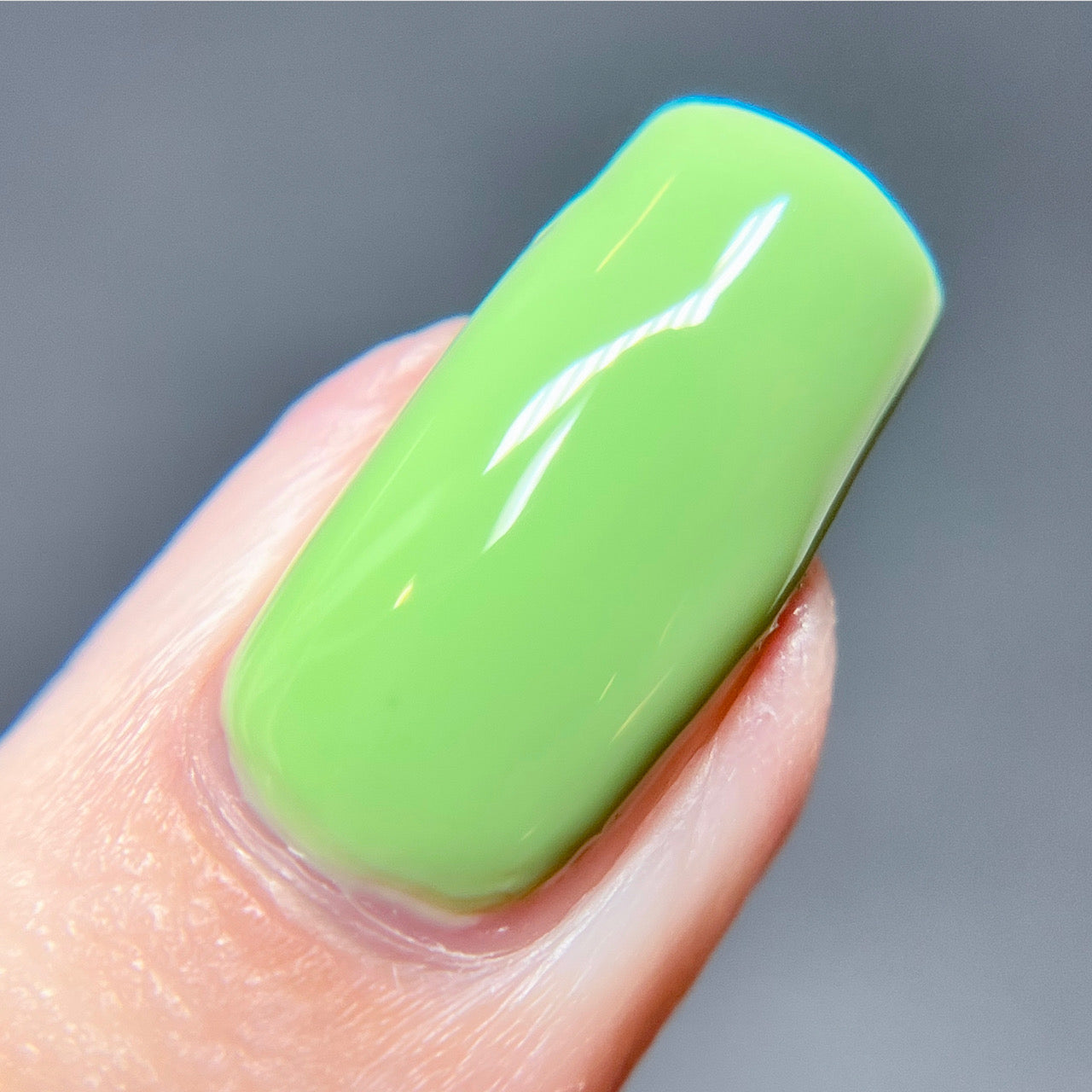 Close-up of a fingernail painted in a glossy, vibrant shade from PI Colors' Pistachio.400 Green Nail Polish line. This exquisite hue captures attention effortlessly with its bright lime green color, reminiscent of pistachios. The background is softly blurred in a neutral gray tone.