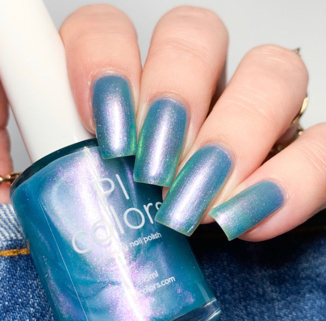 A hand with long, square-shaped nails is adorned with a shimmery blue-green gradient using the handmade Ocean Whitecap.309 Blue Nail Polish from PI Colors. The hand holds a bottle of the same shade, highlighting its color-changing effects in different lighting. A backdrop of denim fabric adds an additional layer of texture to the scene.