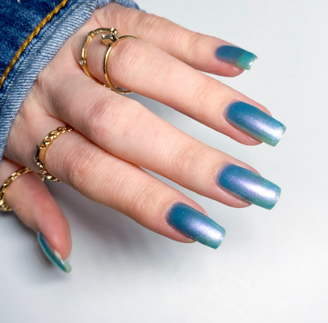 A hand with long, shiny blue-green ombre nails and gold rings on each finger glimmers vibrantly. Adorned in a denim jacket, the nails boast Ocean Whitecap.309 Blue Nail Polish by PI Colors, featuring a handmade formula that delivers an iridescent, color-changing effect that captivates every eye.