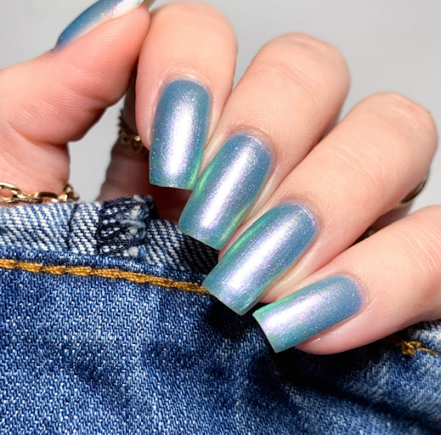 A close-up of a hand with long, glossy nails painted in PI Colors' Ocean Whitecap.309 Blue Nail Polish reveals an enchanting iridescent blue-green hue. The fingers hold the edge of denim fabric with visible yellow stitching, highlighting the stunning color-changing effects that shift gracefully with every movement.