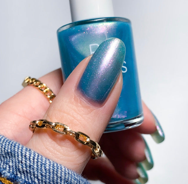 Close-up of a hand holding a bottle of Ocean Whitecap.309 Blue Nail Polish by PI Colors. The nails sport the same shimmering blue-green hue with an alluring pearlescent finish. The person displays gold chain rings, and a denim sleeve is visible, enhancing the vibrant appeal of the handmade nail polish.