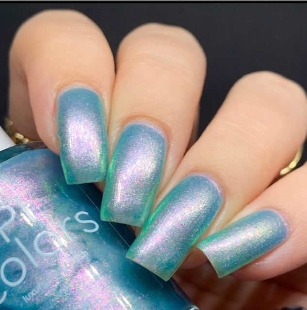 A close-up shot captures a hand with long nails adorned with a shimmering blue-green nail polish, highlighting its enchanting color-changing effects. The individual holds a bottle of the same handmade polish, branded as "Ocean Whitecap.309 Blue Nail Polish" by PI Colors. The dark backdrop enhances the vibrant and mystical hues of the nails.