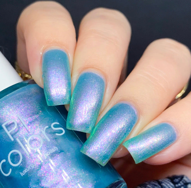 A hand with long, neatly manicured nails is adorned with the enchanting Ocean Whitecap.309 Blue Nail Polish, featuring a mesmerizing blue-to-purple gradient effect. The same handmade polish bottle is held in the hand, proudly displaying the label "PI Colors.