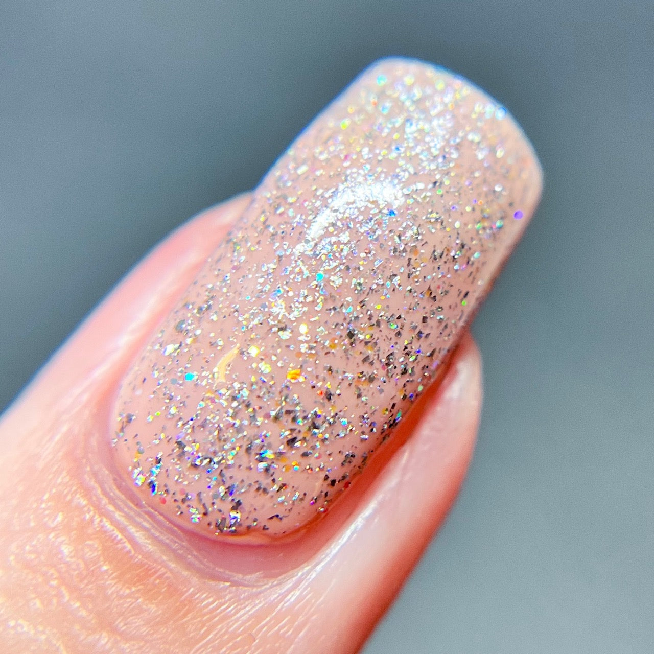 A close-up of a fingernail coated with PI Colors' Diamond Blast.401 Silver Holographic Glitter Nail Polish Topper showcases a translucent pink base sprinkled with fine silver holographic glitter, delivering a dazzling sparkle. This 10-free luxury nail lacquer ensures an elegant finish, complemented by the softly blurred background for added allure.