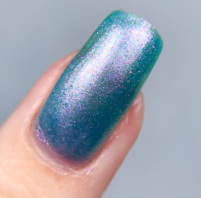 A close-up of a single fingernail features the stunning Ocean Whitecap.309 Blue Nail Polish by PI Colors, showcasing a shimmering gradient that transitions from teal at the base to a purple-tinted tip. Its glossy finish reflects light beautifully, enhancing the sparkly texture and color-changing effects of this blue-green masterpiece.