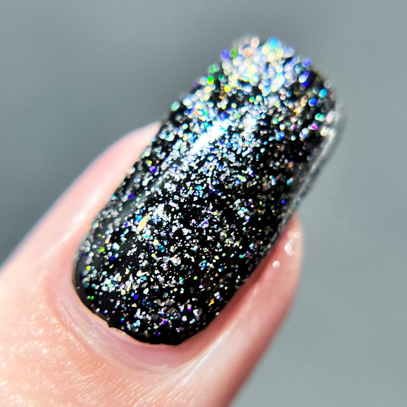 A close-up showcases a fingernail coated in black nail polish with PI Colors' Diamond Blast.401 Silver Holographic Glitter Nail Polish Topper layered on top. The holographic sparkles catch the light, producing a shimmering effect. The blurred background highlights the textured and glossy finish of this exquisite nail art.