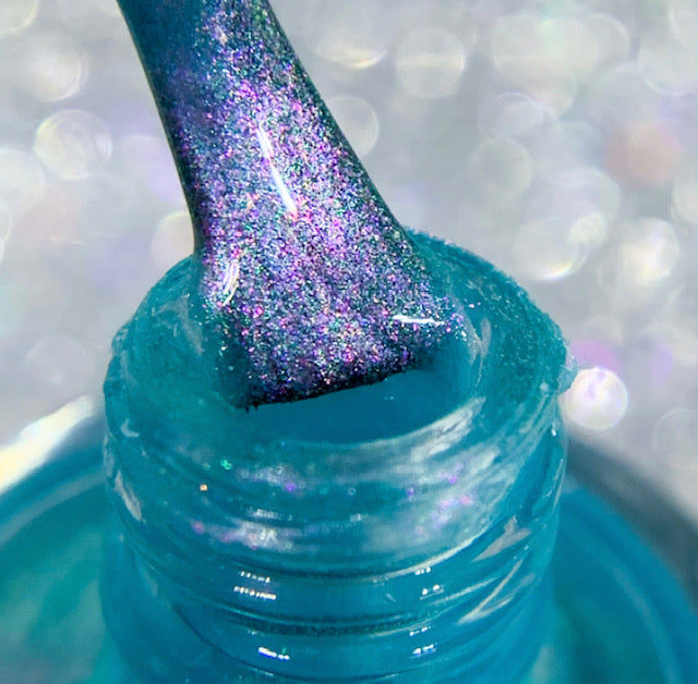 A close-up shows a PI Colors Ocean Whitecap.309 Blue Nail Polish bottle, with a shimmering blue brush being pulled out. This nail polish features a sparkling texture with hints of purple and enchanting color-changing effects. The softly blurred background enhances the bokeh effect.