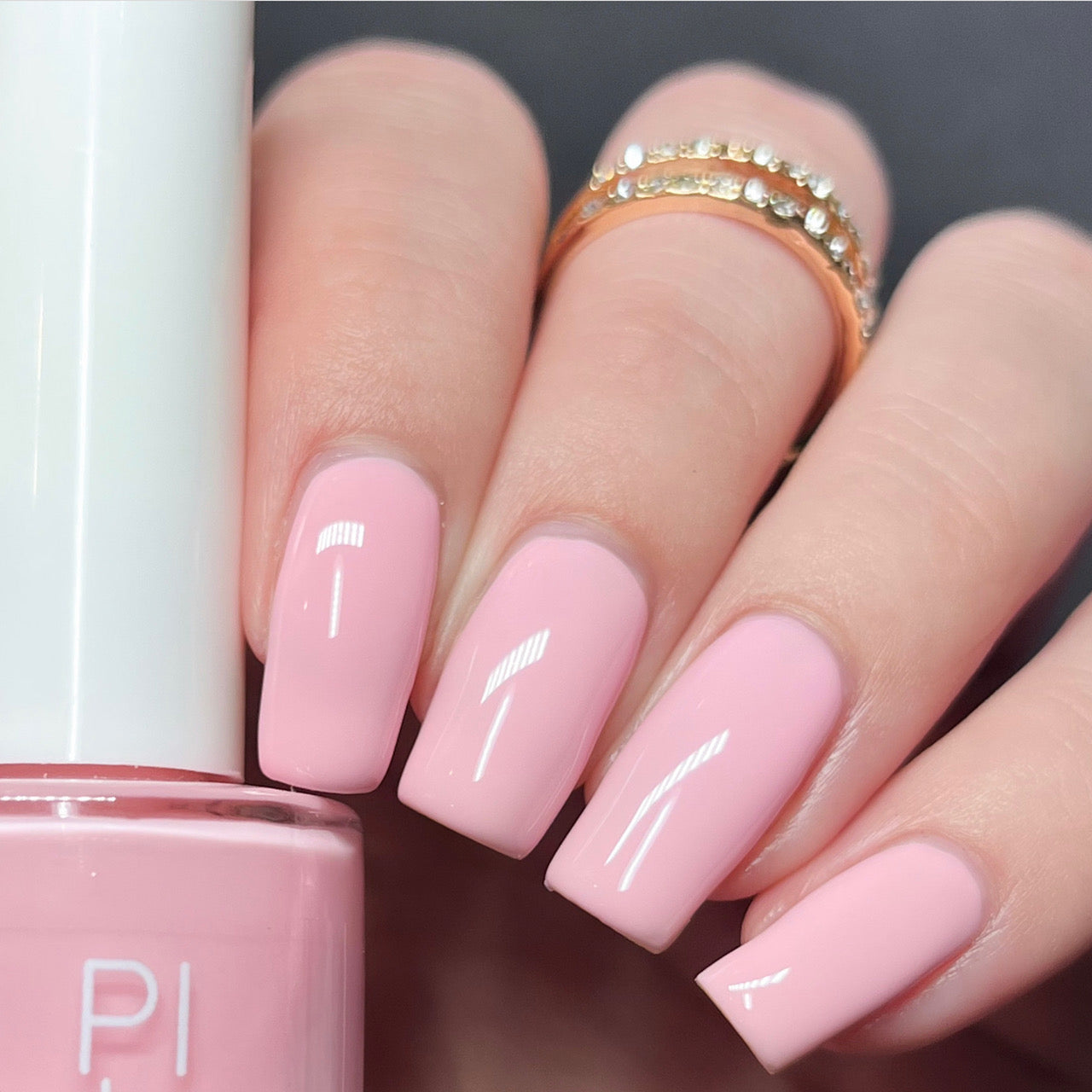 A hand featuring medium-length, square-shaped nails adorned with a glossy, pastel pink from the luxurious Cupcake.400 Pink Nail Polish by PI Colors. The person is wearing a delicate gold ring embedded with small diamonds and is holding a pink nail polish bottle marked "PI," suggesting its exquisite creme finish, between the thumb and index finger.