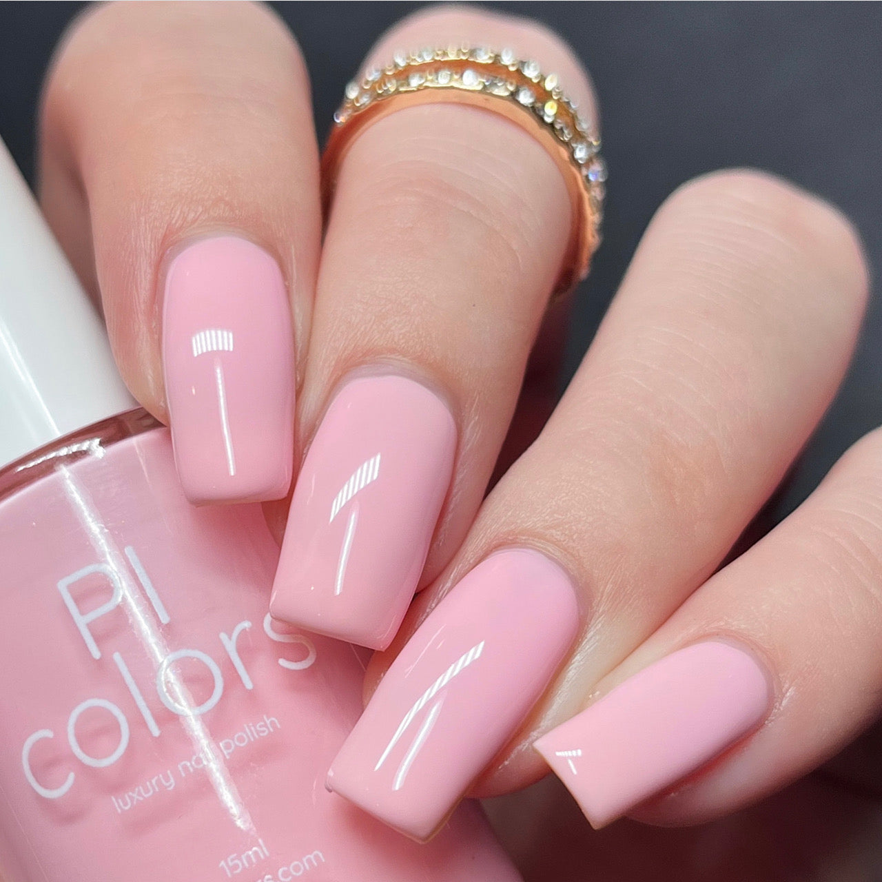 A close-up of a hand with long, square-shaped nails painted in the glossy pastel pink shade of Cupcake.400 Pink Nail Polish. The person is wearing a gold ring adorned with small diamonds on the ring finger. A bottle from PI Colors is held in the hand, showcasing its exquisite creme finish.