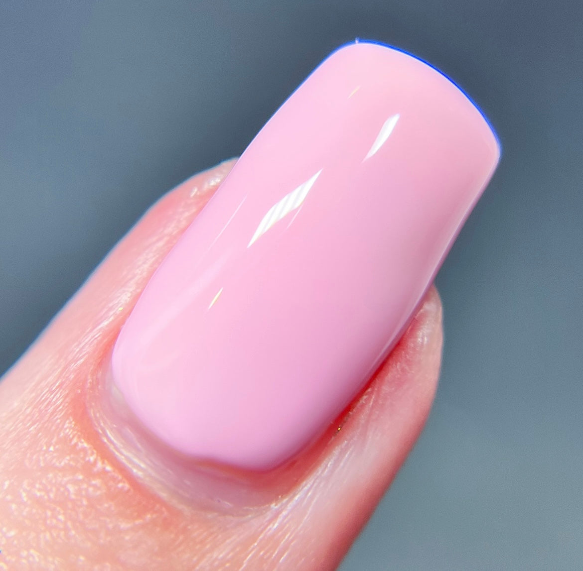 A close-up shot of a fingernail adorned with Cupcake.400 Pink Nail Polish by PI Colors, featuring a smooth, pastel pink creme finish. The background is softly blurred in shades of blue and gray.