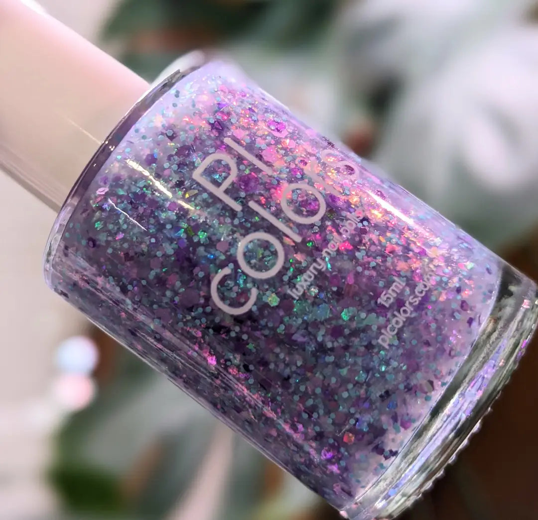 A close-up of a "Hyacinth Rains.000" nail polish bottle from PI Colors sparkles with a 10-free formulation and features colorshifting rainbow flakies within its dazzling blend of purple, blue, and pink glitter. The blurred background subtly reveals touches of green and brown.