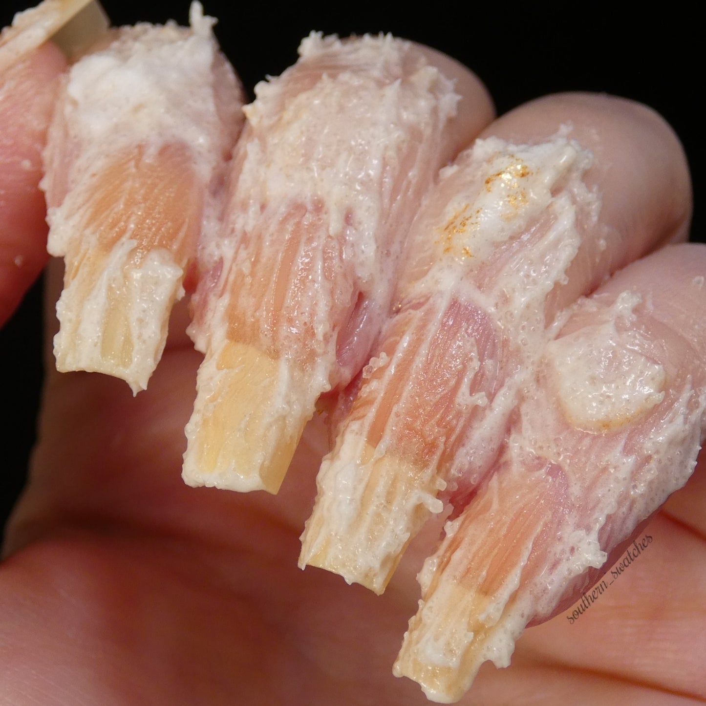 Fingers with long nails are coated in a thick, textured layer of PI Colors Honey Scented Whipped Soap Sugar Scrub, resembling intricate patterns of whipped soap. The dark background enhances the striking contrast, as subtle hints of honey scent linger in the air.