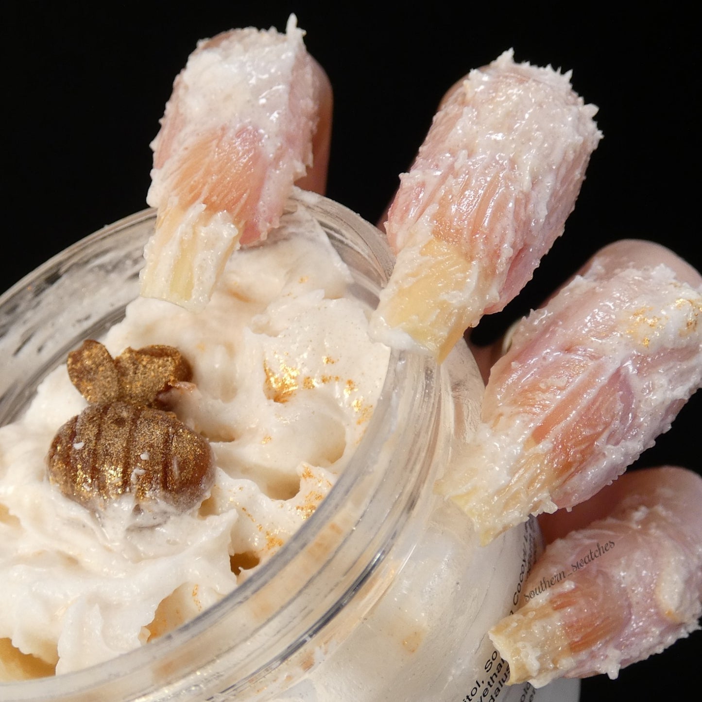 A pair of human fingers with nails styled to resemble pink crab claws grasps a jar of PI Colors' Honey Scented Whipped Soap Sugar Scrub. The cream is adorned with a small, gold, pumpkin-shaped ornament. The honey-scented contents pop against the black background, emphasizing the distinctive nail art and the jar's charm.