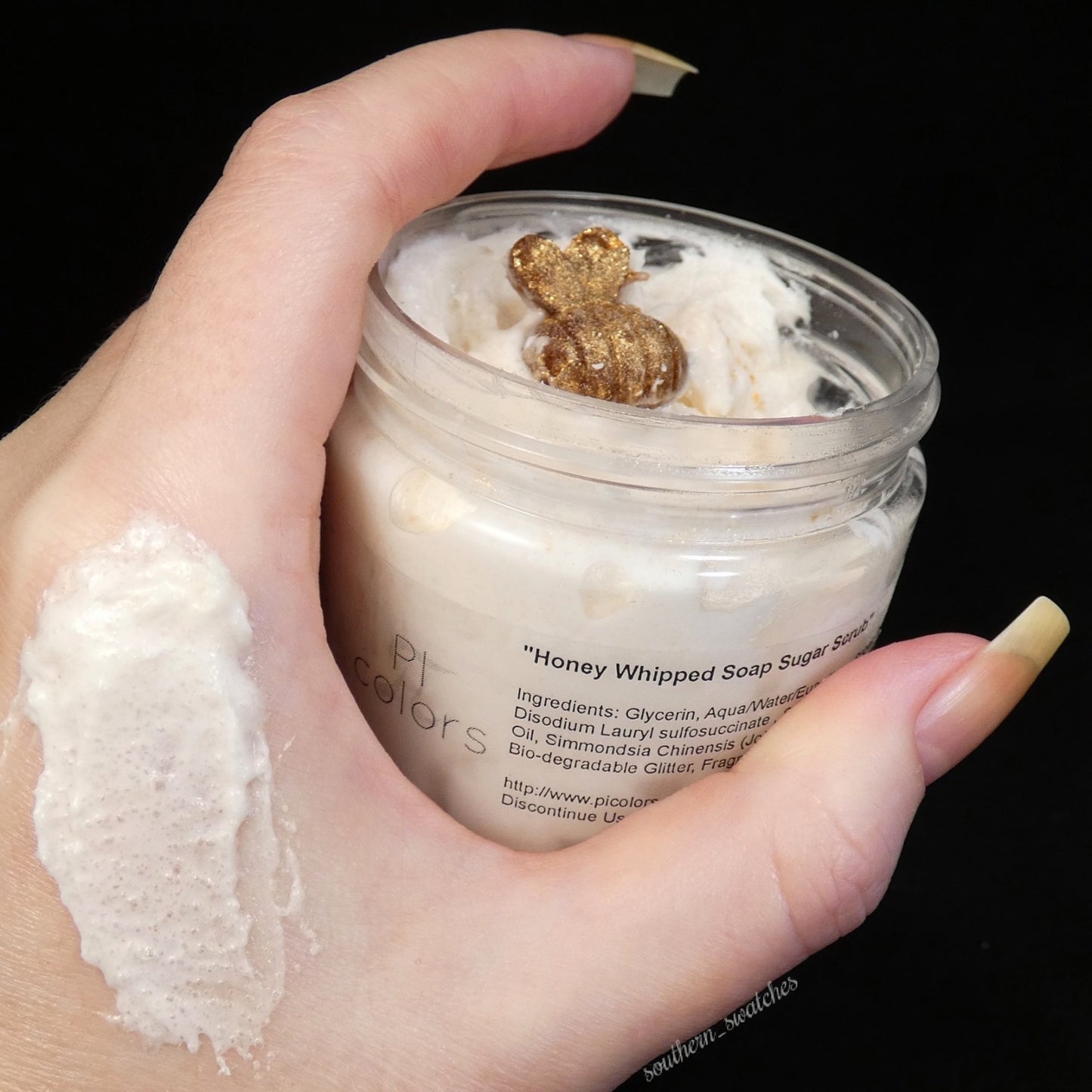 A hand showcases an open jar of the "Honey Scented Whipped Soap Sugar Scrub" by PI Colors, featuring a delightful honey fragrance and adorned with a charming decorative bear on top. The ingredients are displayed on the label, while a swatch of the rich scrub is applied to the back of the hand.