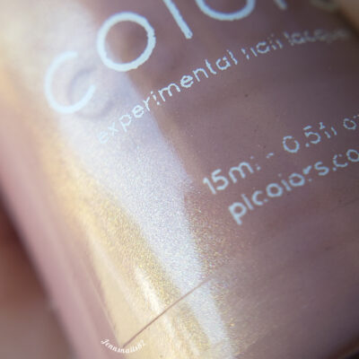 Close-up of a luxury nail lacquer bottle from PI Colors, showcasing its iridescent, light-reflecting surface. The label reads "Milk Berry.073 Pink Nail Polish," indicating it's a handmade polish, and it also displays volume and website information. The image highlights the shimmery texture of this exquisite polish.