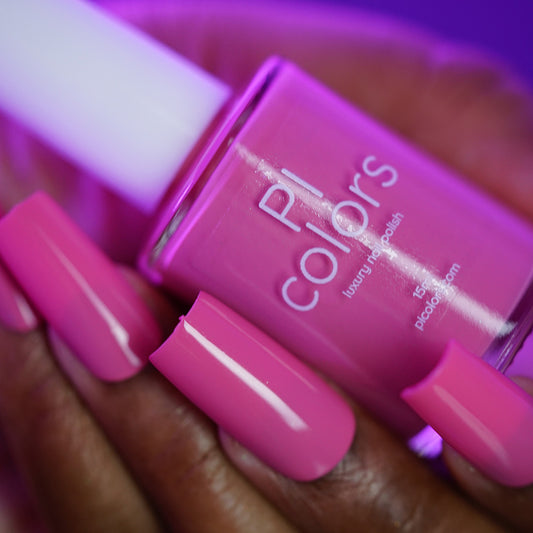 A hand with long, glossy, bright pink nails holds a PI Colors Neapolitan.432 nail polish bottle with a white cap, showcasing the vibrant shade of this luxury lacquer.