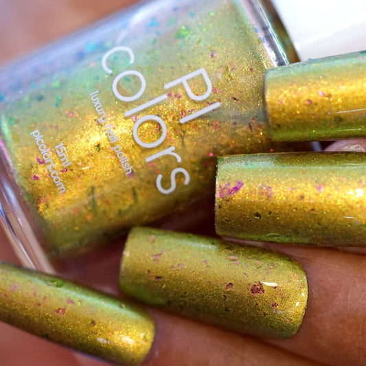 Close-up of manicured nails painted with shimmery green nail polish, accented with multichrome flakies for an iridescent touch. A bottle of That Golden Ring.000 Gold Nail Polish from PI Colors is in the background, partially visible.