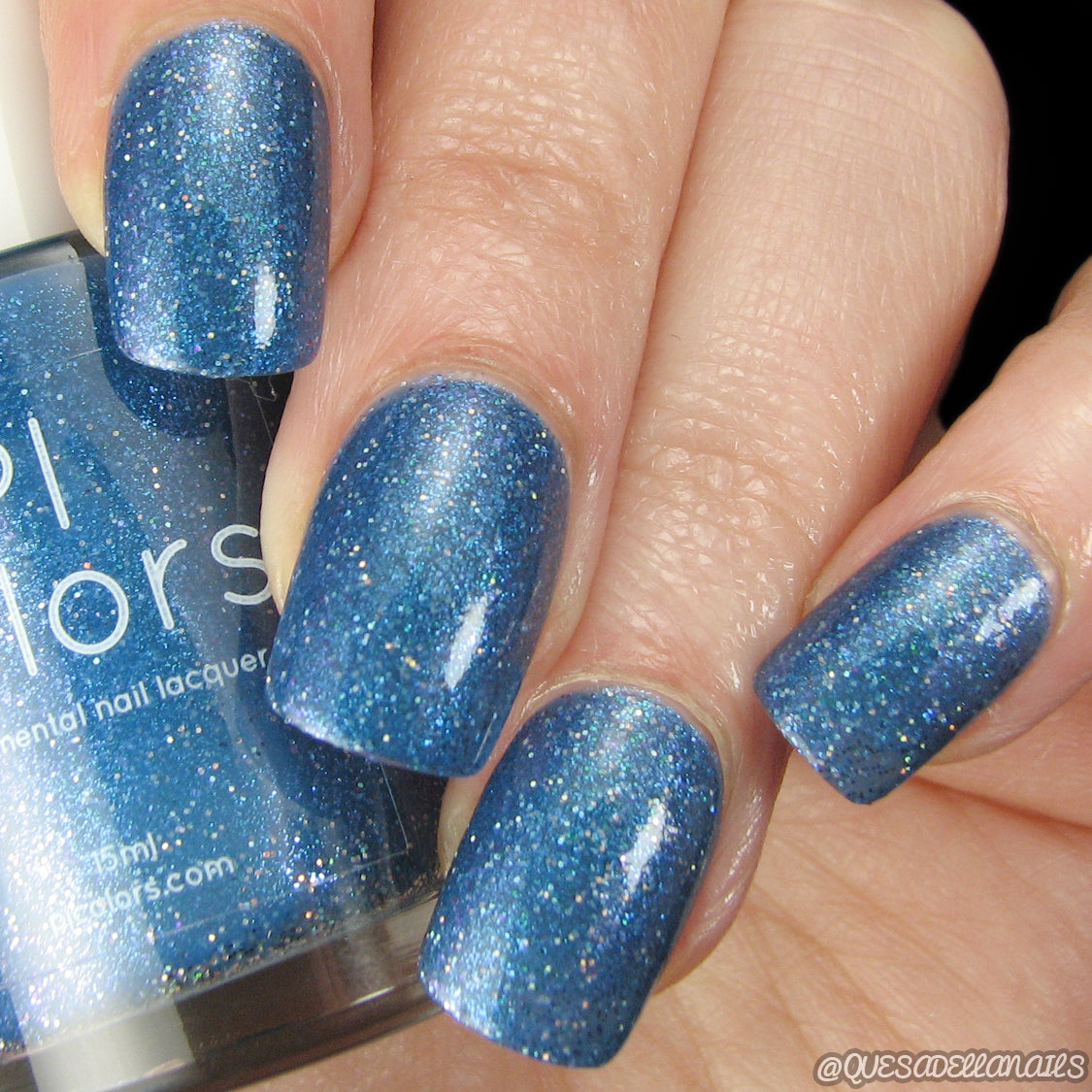 A hand with neatly manicured nails, painted in the shimmering blue shade of Cockatoo.036 from PI Colors, holds a bottle of the same glittery 10-free nail polish. The light reflects off the dazzling finish, enhancing its luxurious appeal. A simple backdrop keeps all attention on the nails and bottle.
