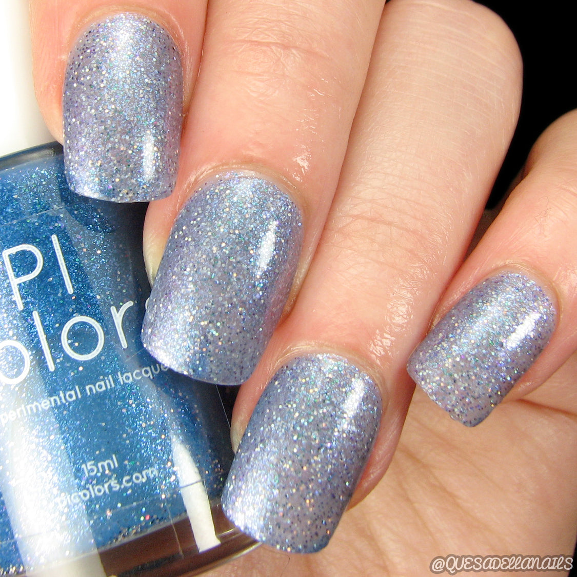 Close-up of a hand with nails painted in PI Colors' Cockatoo.036 Blue Nail Polish, showcasing a glittery blue-purple finish. The 10-free nail polish bottle, partially visible, matches the nail color perfectly and offers a textured, sparkling finish that's both elegant and vibrant.
