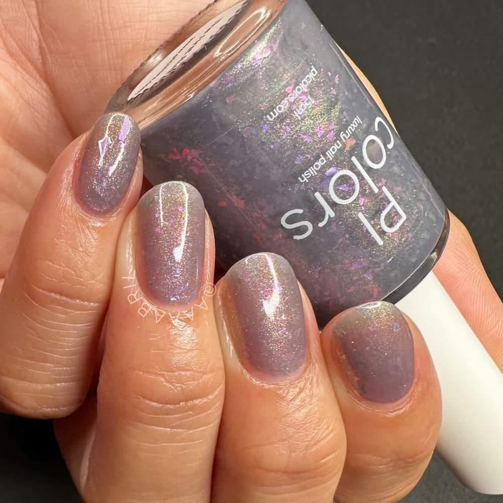 A close-up of a hand with short, neatly manicured nails displays the shimmery grayish-purple hue of "The First Rule.000 Gray Nail Polish" by PI Colors, featuring iridescent flakies. The hand holds a small bottle of this nail polish, crafted in custom batches to match.